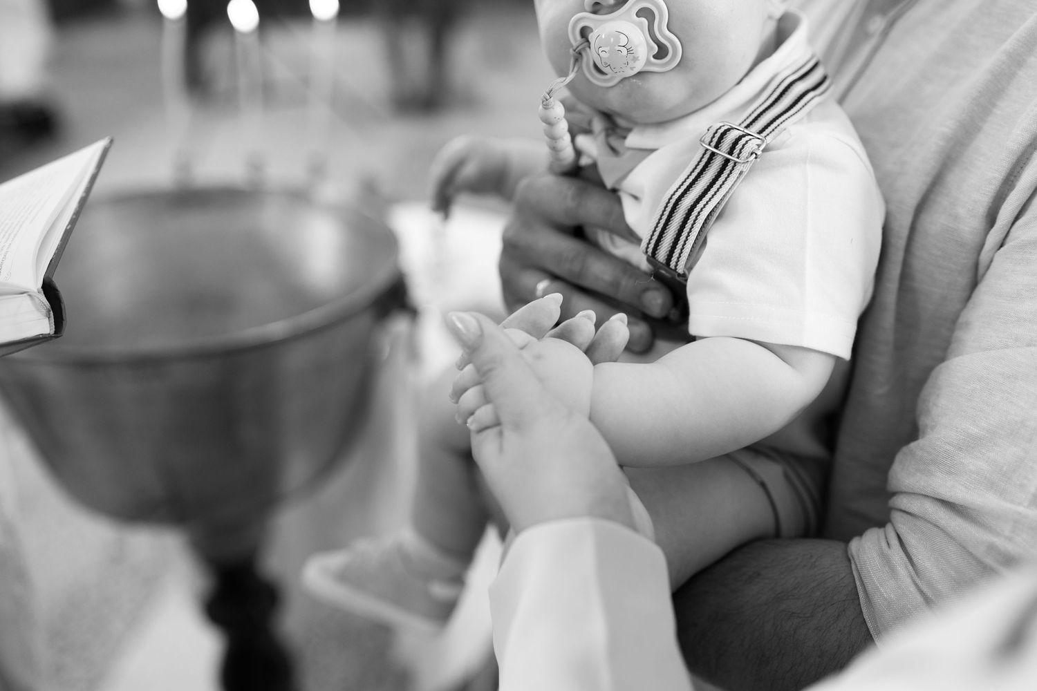 Black and white photo of a baby with pacifier being held during a baptism ceremony near a font.