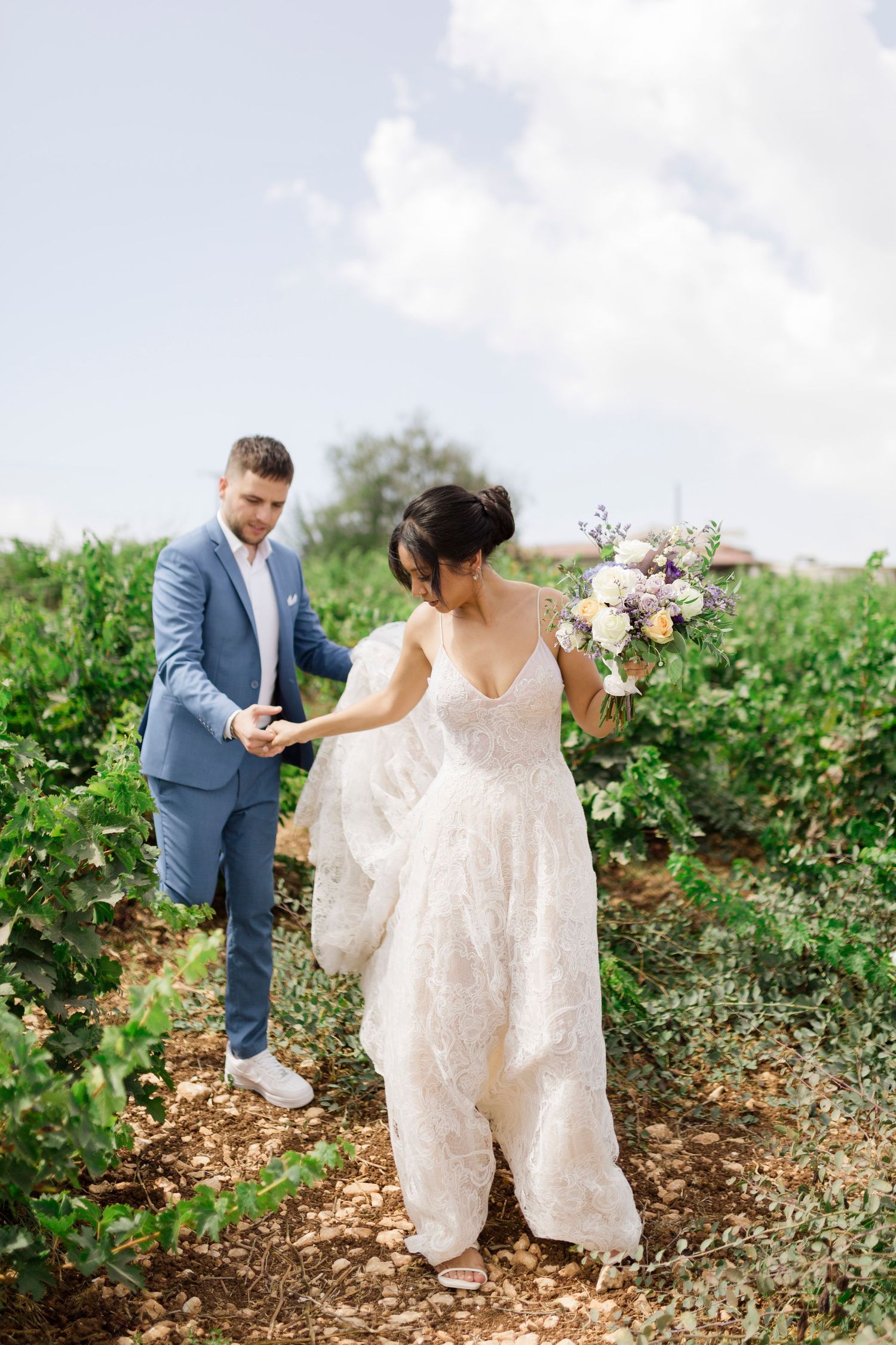 Bride in lace gown holds bouquet as groom in blue suit guides her through lush vineyard on wedding day.
