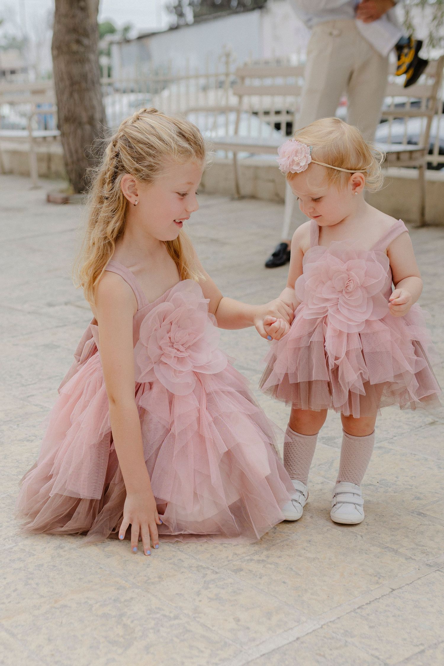 Two young girls in matching pink tulle dresses hold hands at an outdoor event, with the older girl smiling warmly.