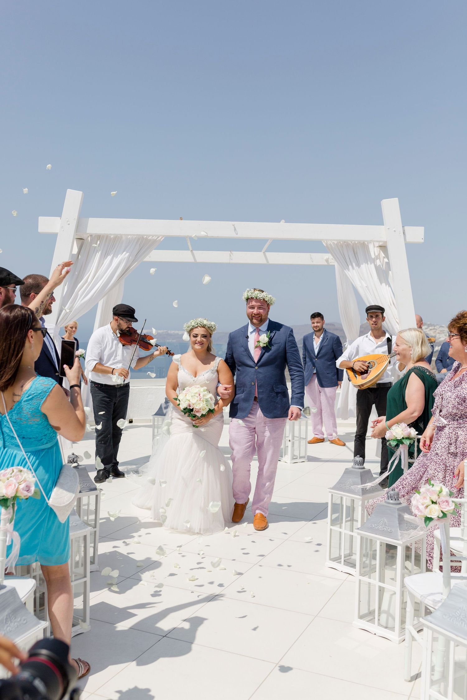 Newlyweds walking down aisle as guests toss petals at outdoor seaside wedding ceremony under white pergola.