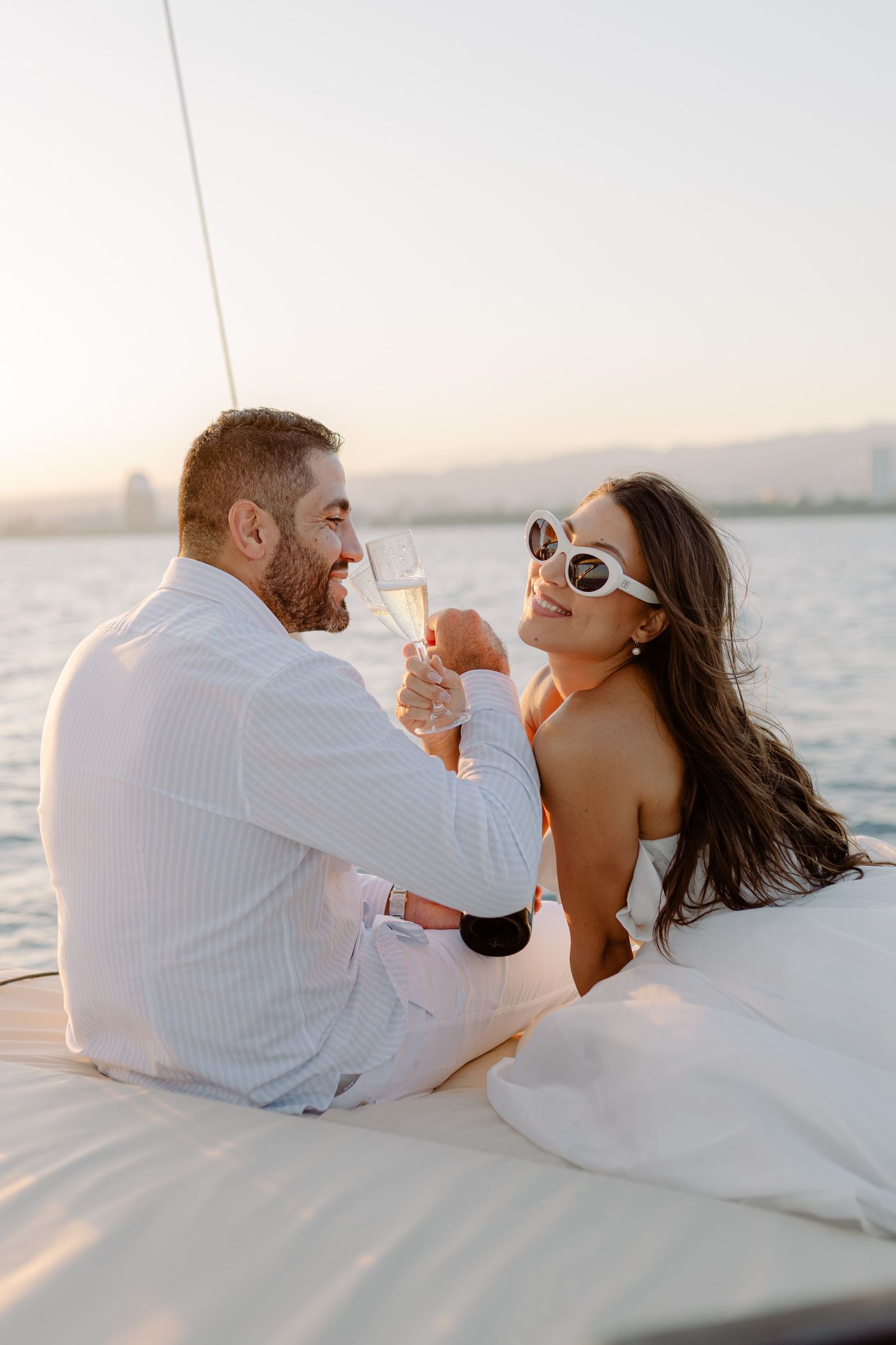 A couple toasting champagne glasses on a luxury yacht at sunset, woman wearing white sunglasses and strapless top.