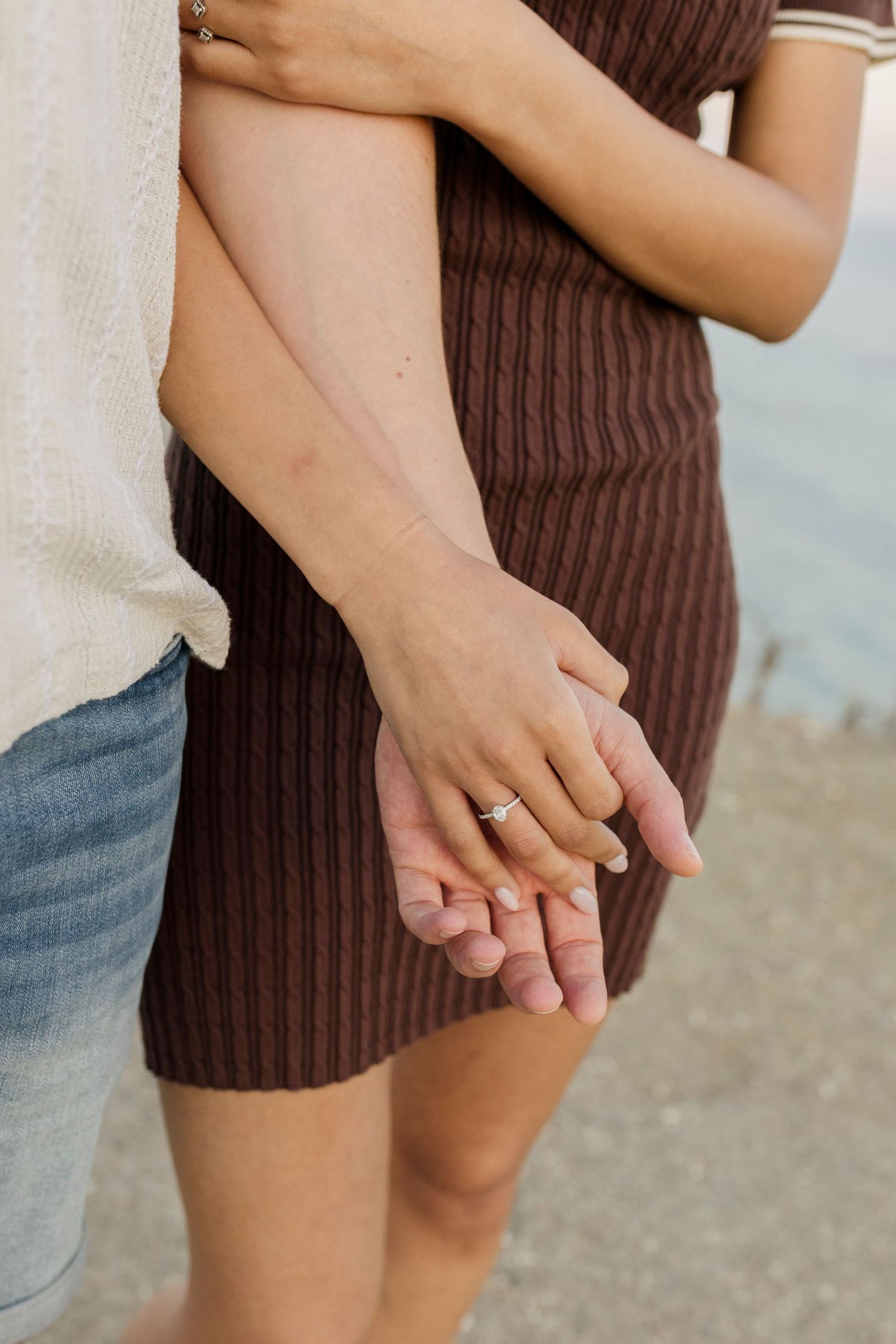 Close-up of couple holding hands, woman wearing engagement ring, man in denim shorts, woman in brown ribbed dress.