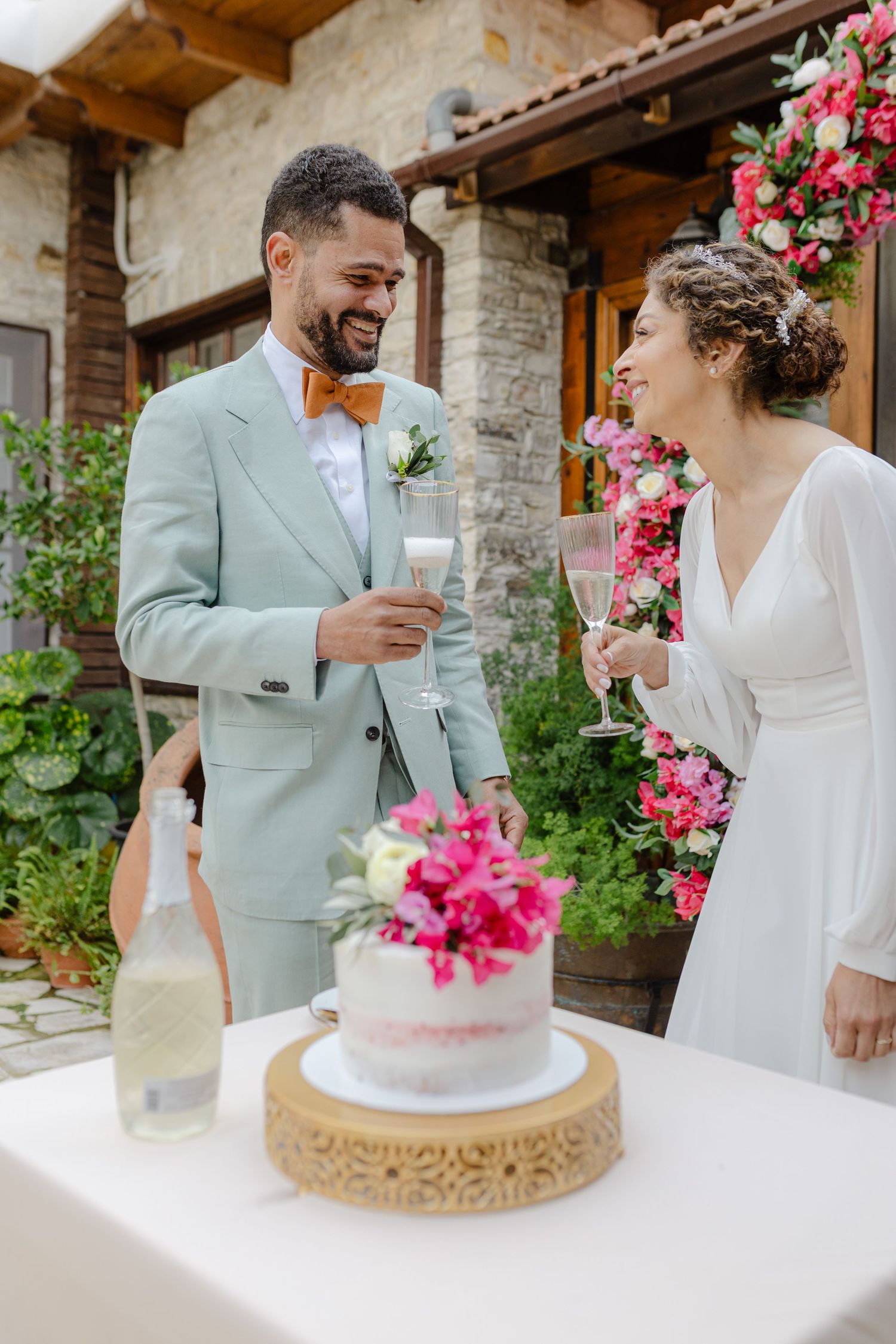Bride and groom toasting with champagne glasses beside a floral wedding cake at an outdoor rustic venue.