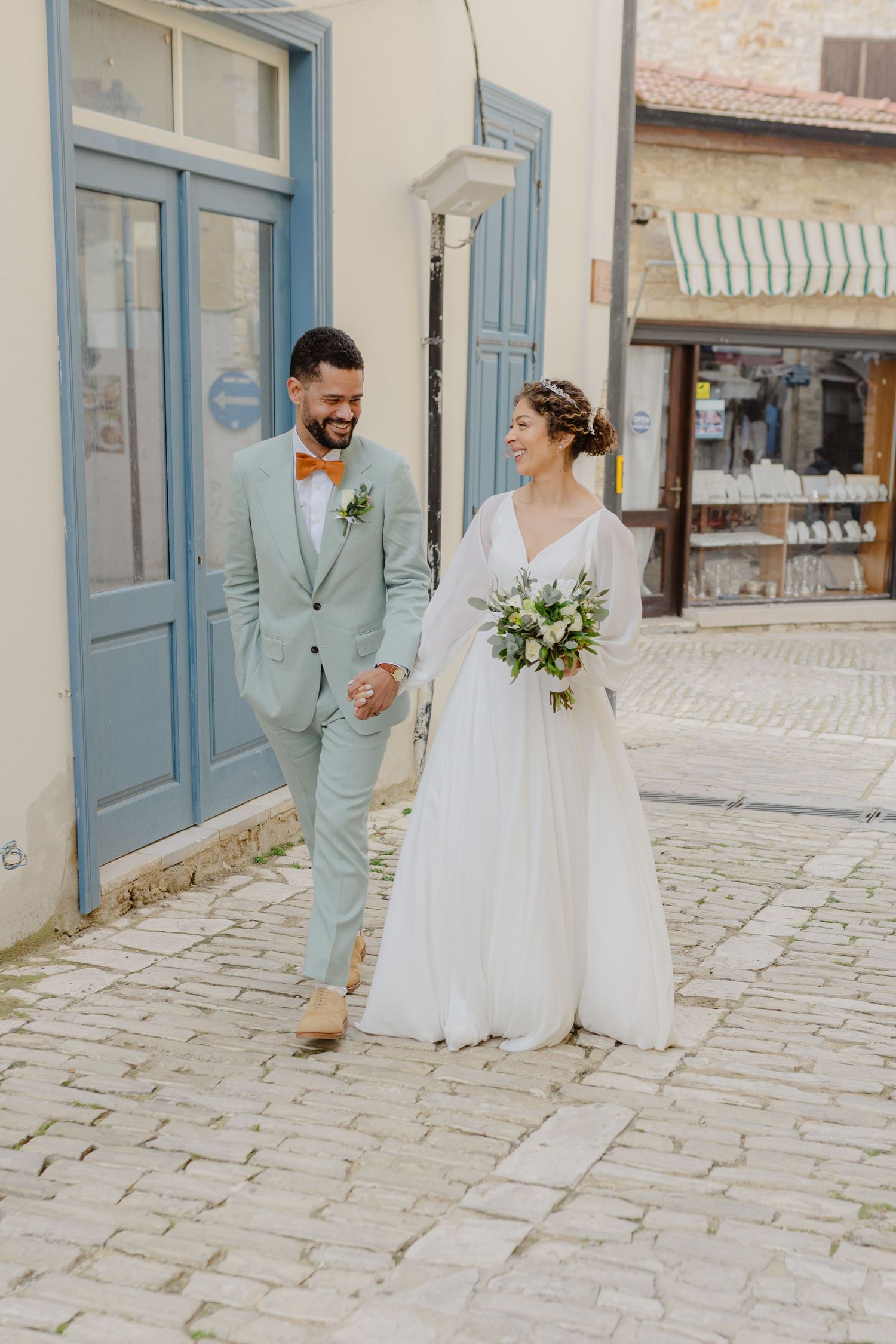 Bride and groom walking hand in hand on cobblestone street, groom in mint suit with orange bow tie, bride in white gown.