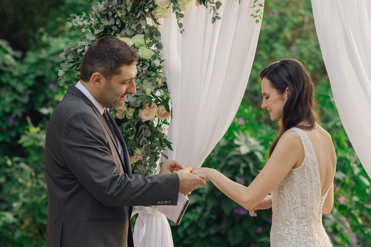 Groom placing wedding ring on bride's finger during outdoor ceremony with floral arch backdrop.