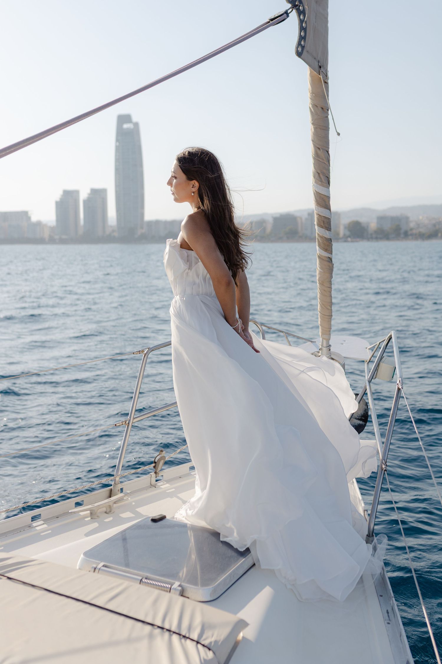 A bride in a flowing white gown stands on a yacht bow, gazing at a city skyline across calm blue waters.