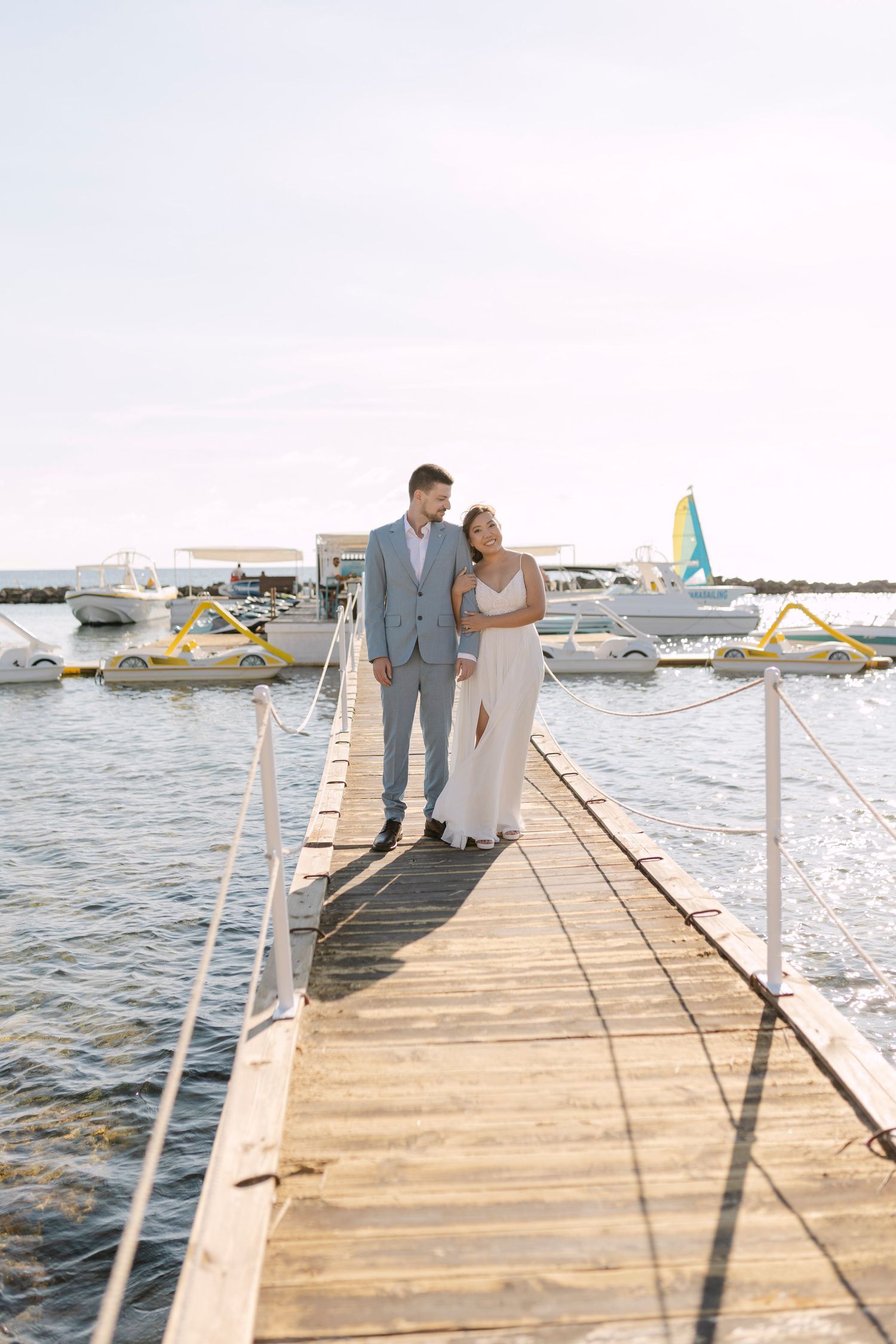 Bride and groom walking hand in hand on a wooden pier with boats and sparkling water in the background.