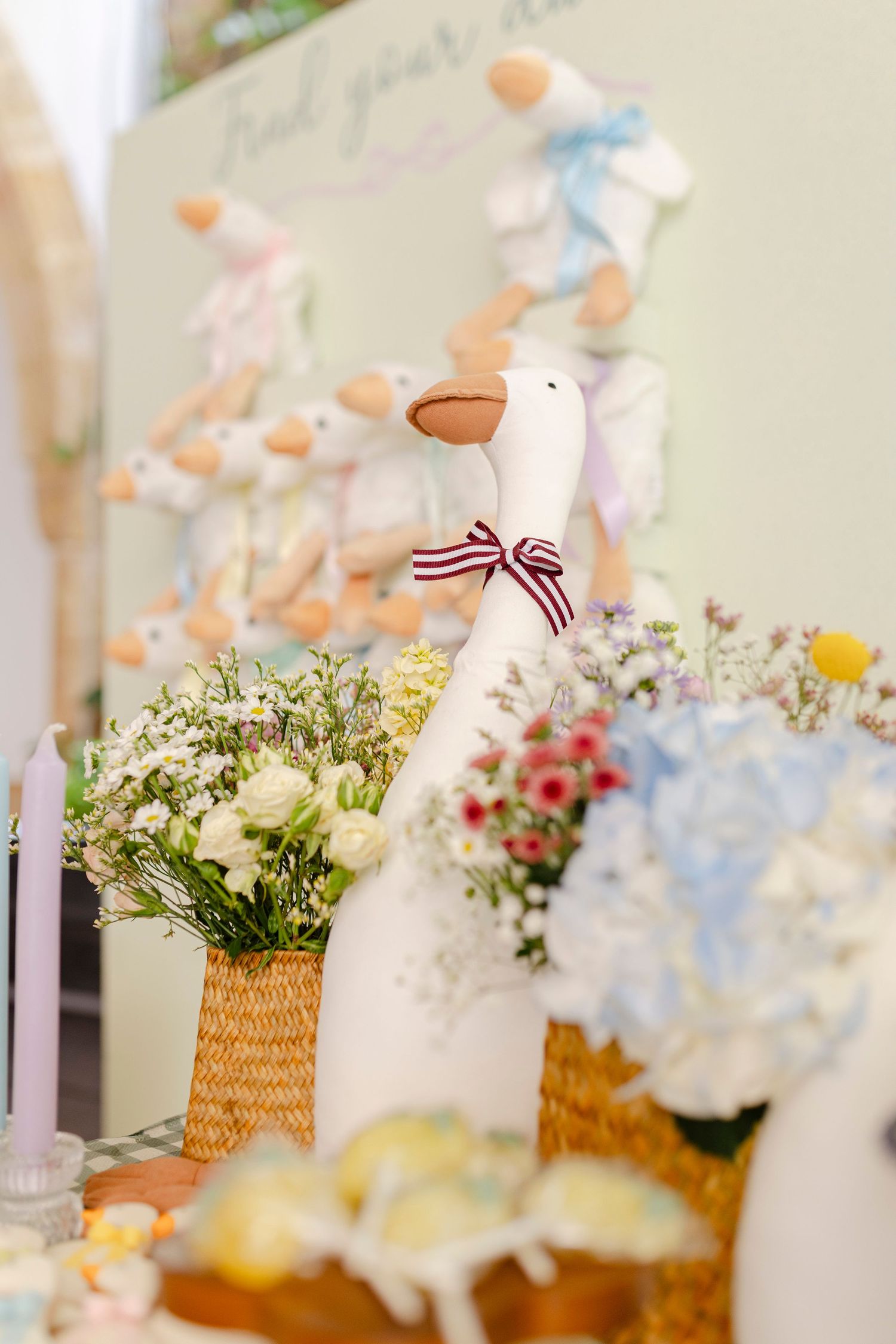 White ceramic goose figurine with striped bow surrounded by colorful flower arrangements and pastel stuffed animal toys on display table.