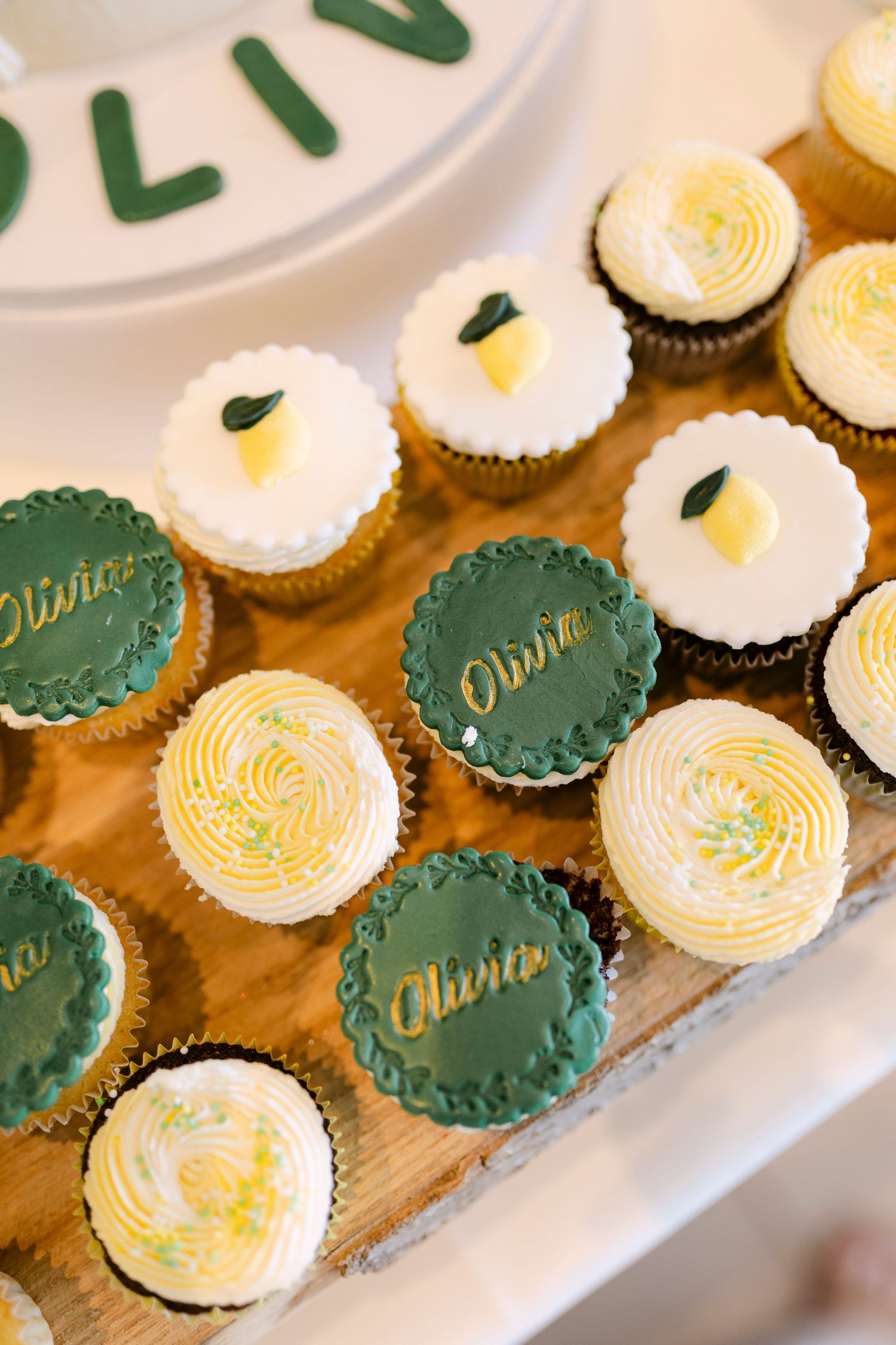 Elegant baptism cupcakes with green fondant toppers inscribed Olivia and lemon decorations on wooden tray.