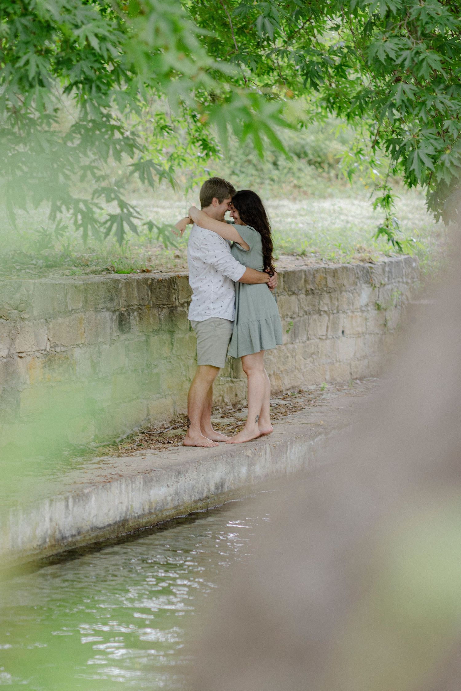 A barefoot couple shares a romantic embrace on a stone ledge near water, surrounded by lush green trees.