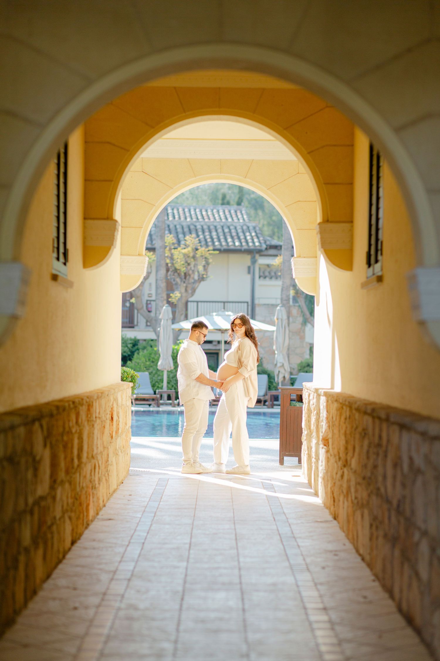 Couple in white outfits embracing under a yellow archway corridor with a pool and resort in the background.