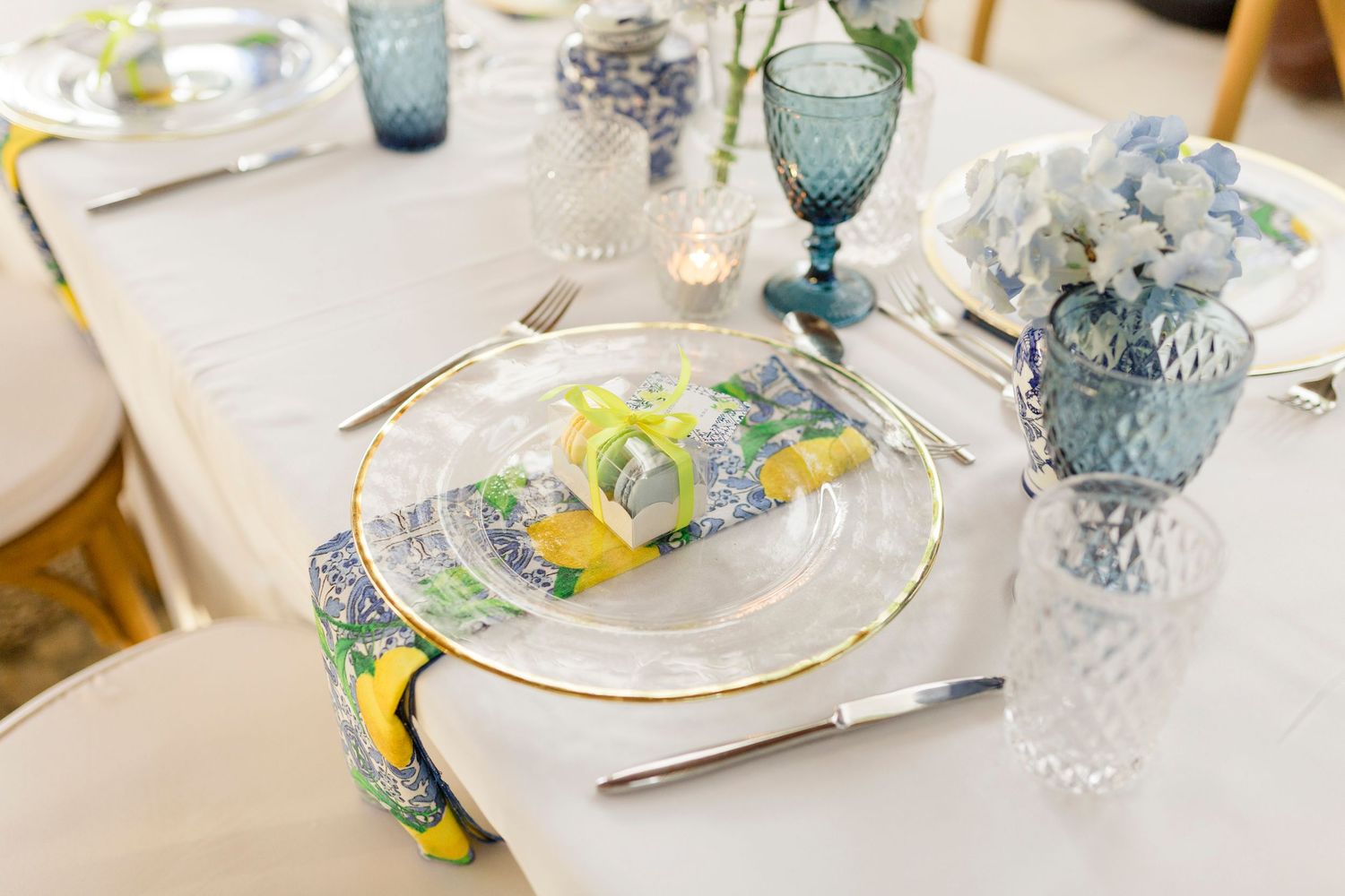 Elegant table setting with gold-rimmed plates, yellow floral napkins, blue glassware, and white hydrangeas centerpiece.