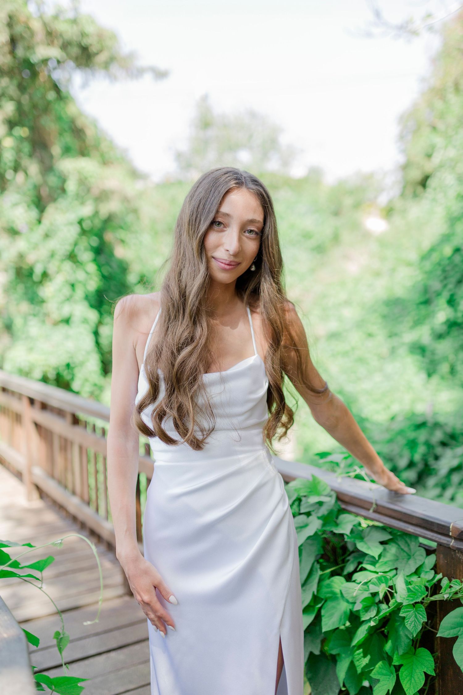 Woman in white satin slip dress poses on wooden bridge surrounded by lush green foliage.