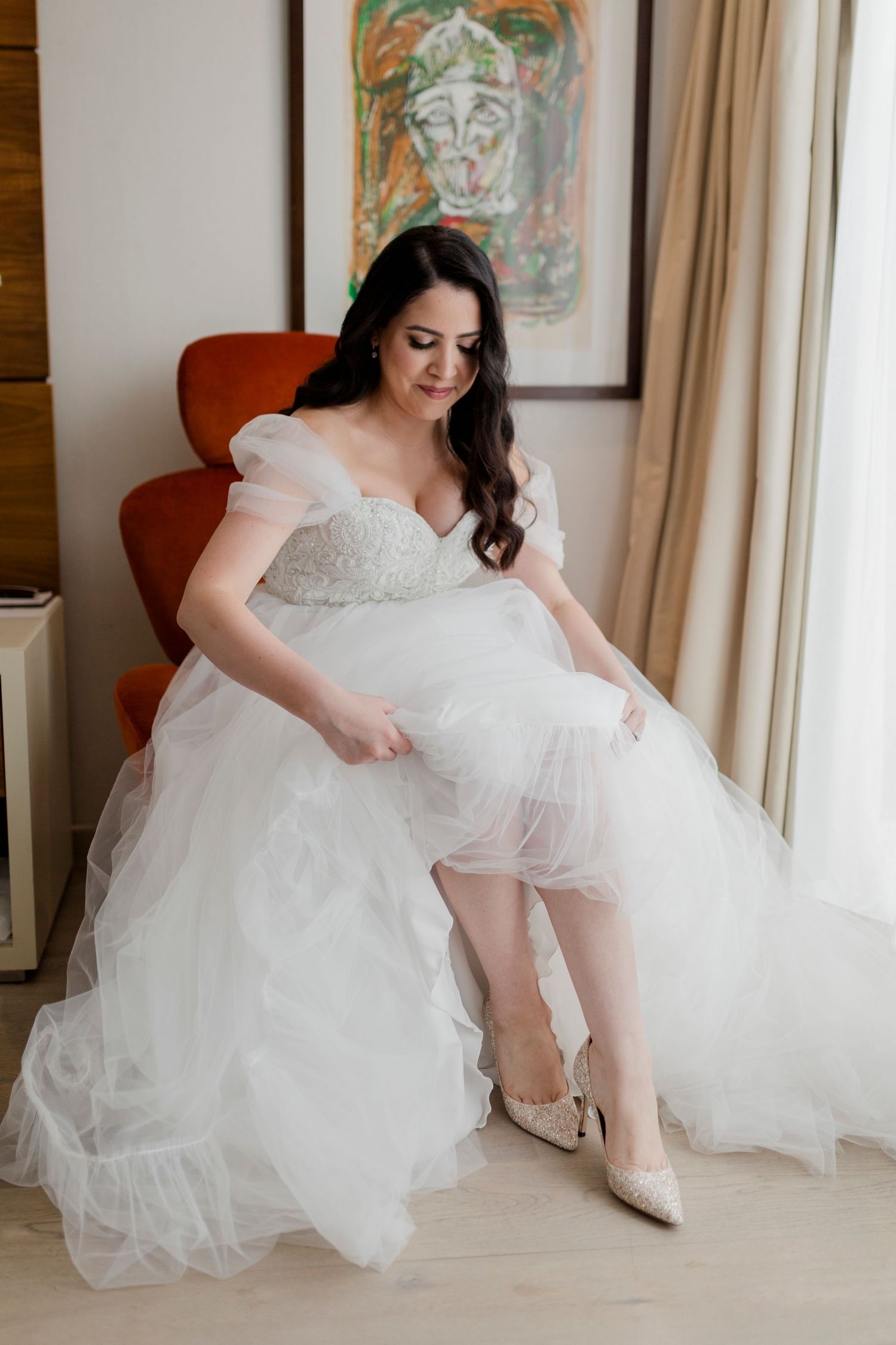 Bride in white tulle ball gown adjusting her heel while seated in a red chair near a bright window.