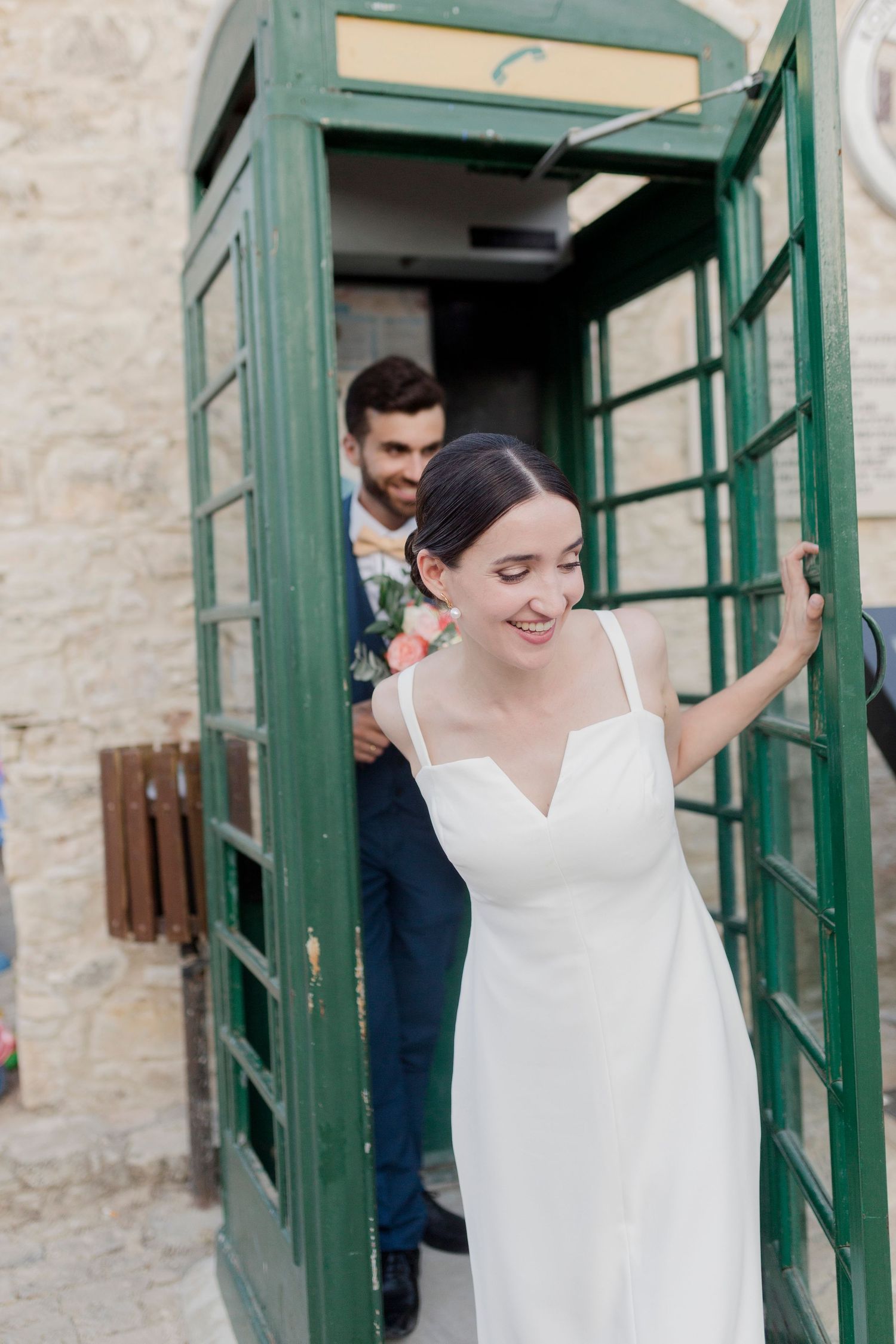 Bride in white spaghetti strap gown stepping out of a green vintage phone booth with groom.