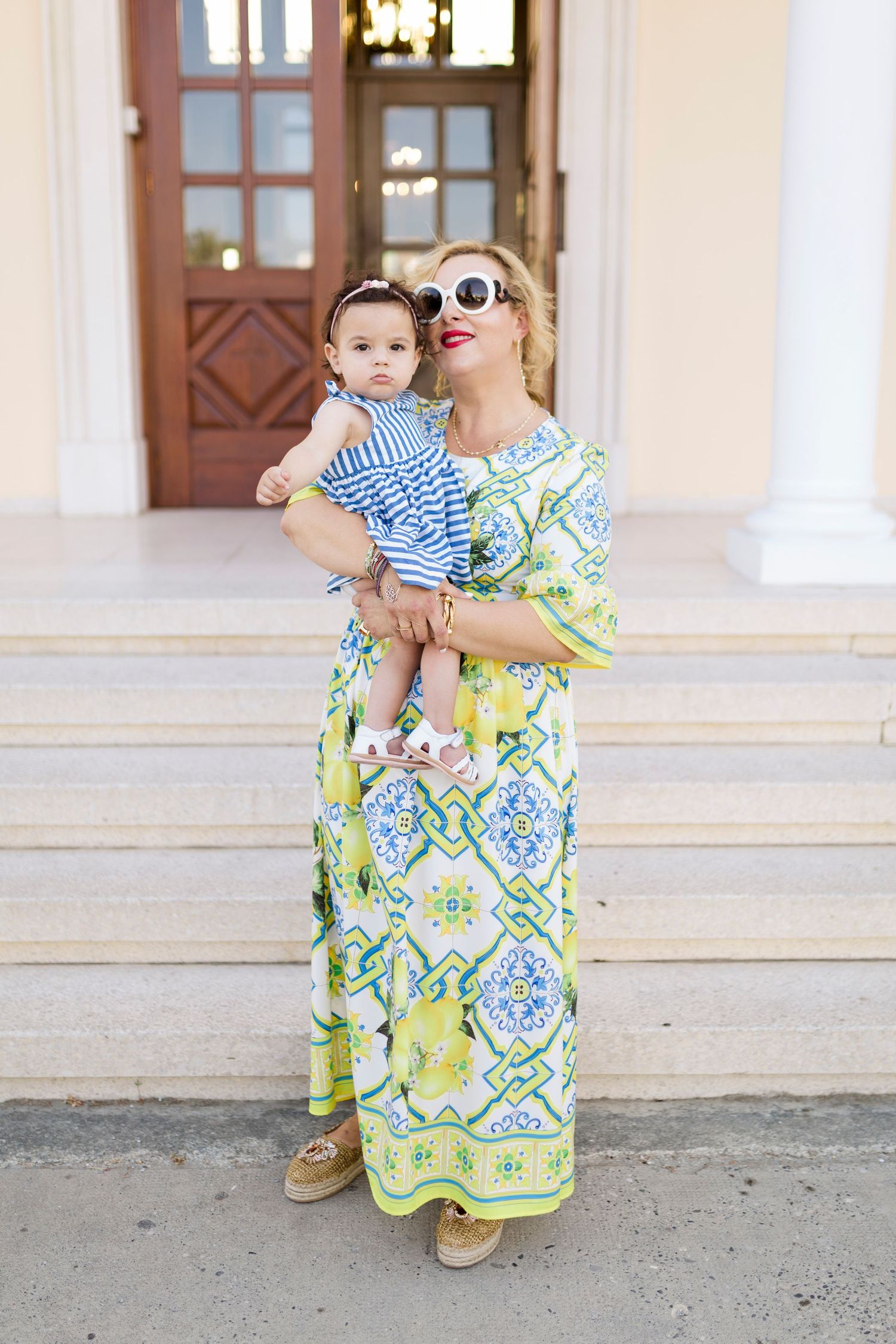 Woman in colorful maxi dress holding baby in striped outfit on building steps.