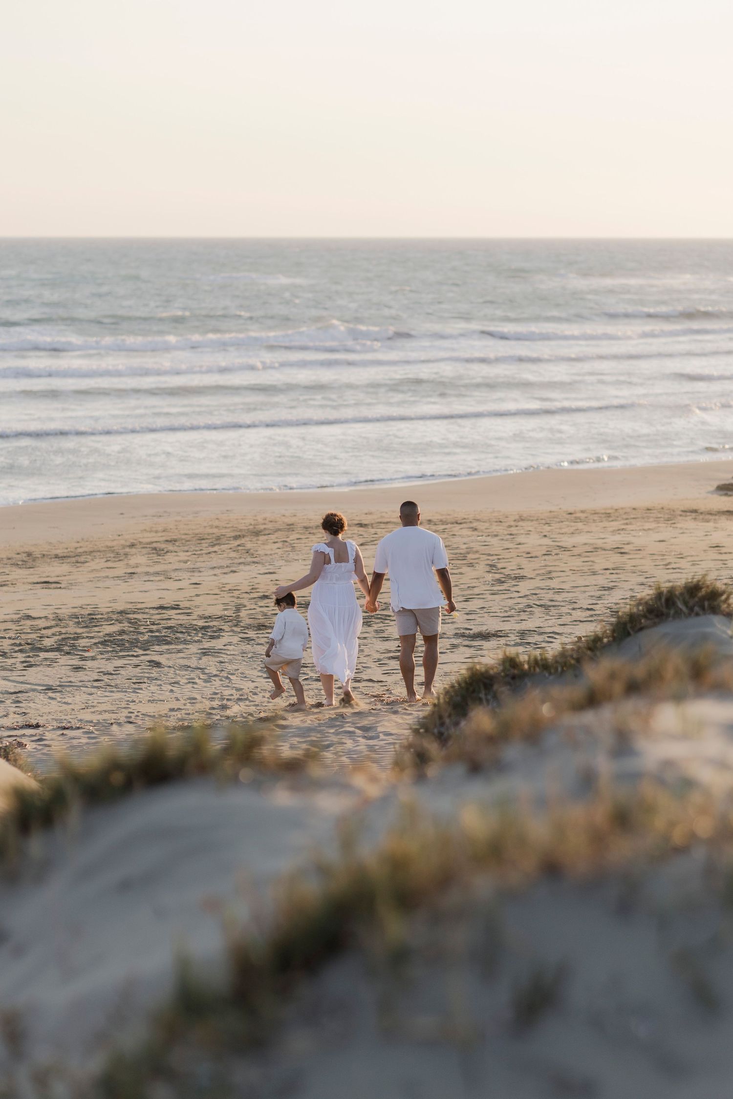 Family in white outfits walking hand-in-hand toward ocean waves on a sandy beach at sunset.