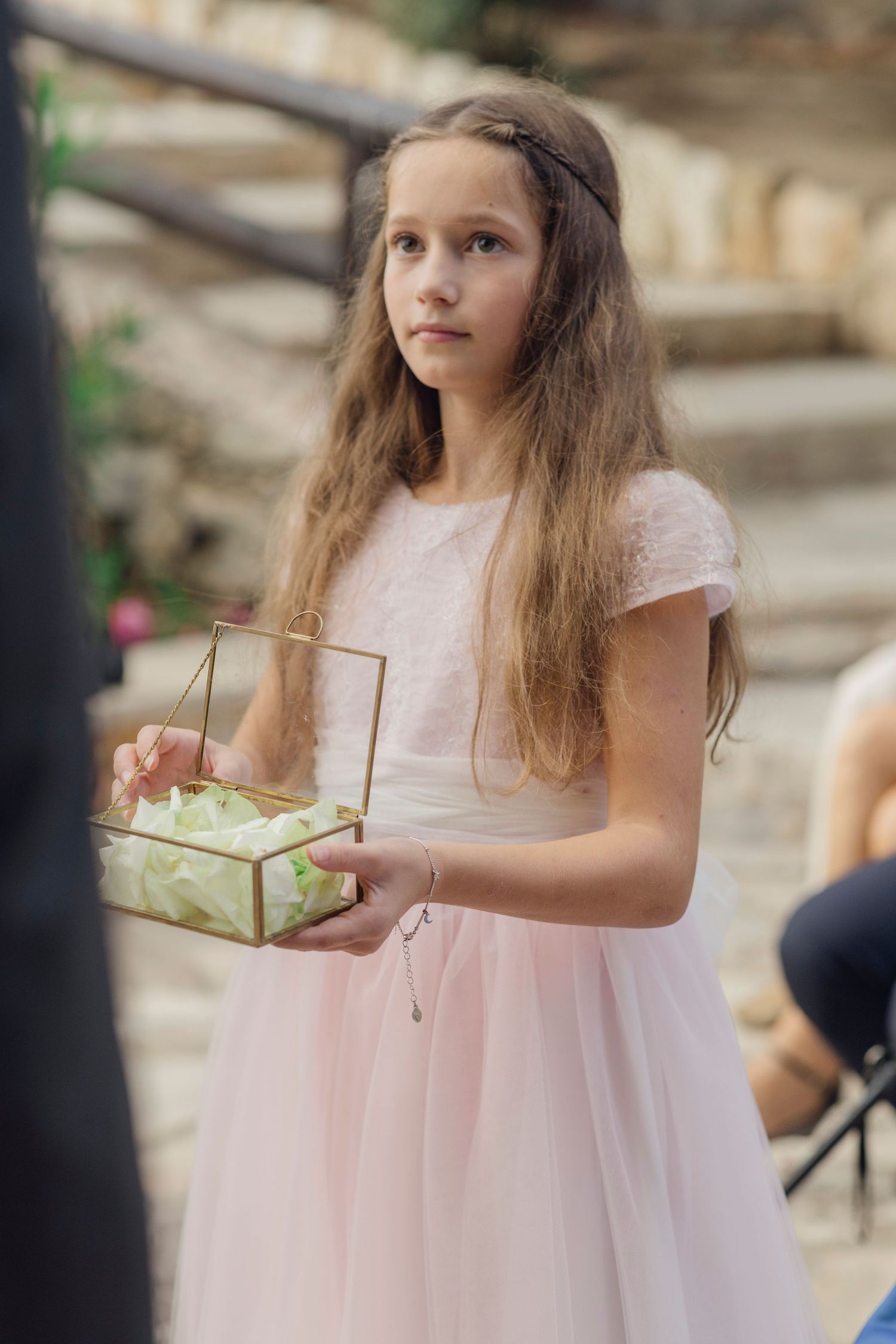 Young flower girl in pink dress holds glass ring box at outdoor wedding ceremony.