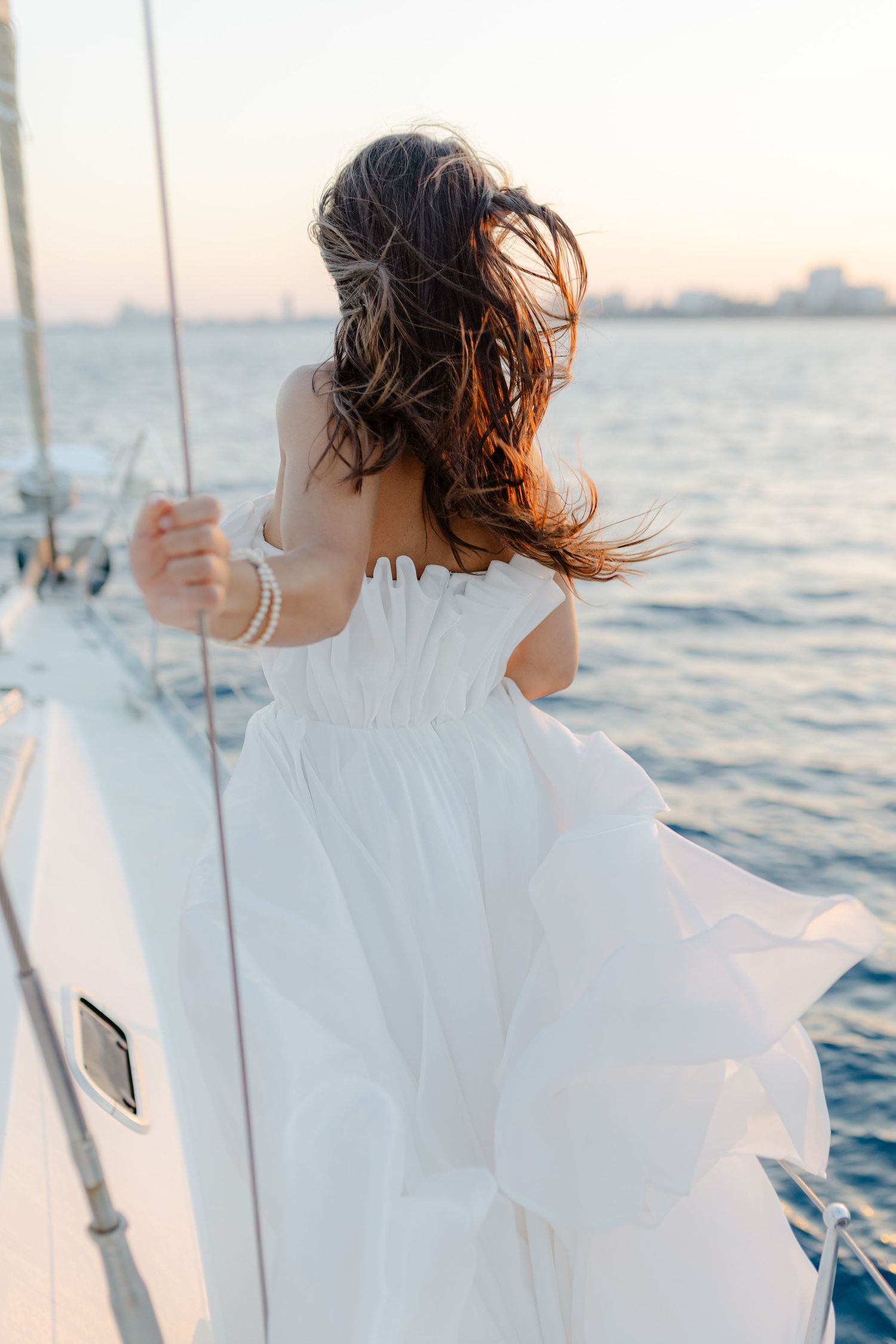 Bride in white strapless gown standing at sailboat railing with hair blowing in wind.