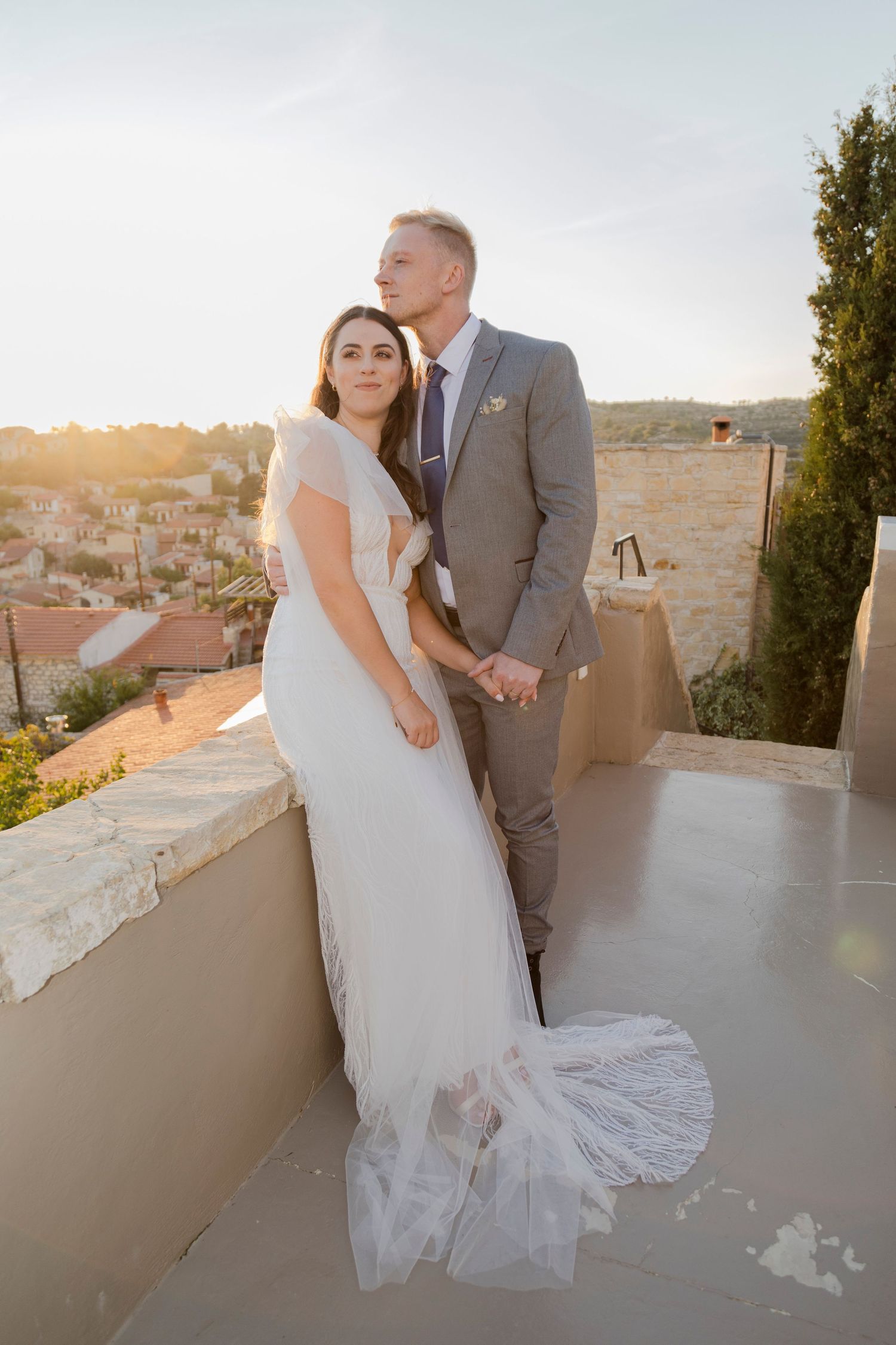 Bride and groom posing on a rooftop terrace at sunset, with a scenic village backdrop, bride in white gown, groom in grey suit.