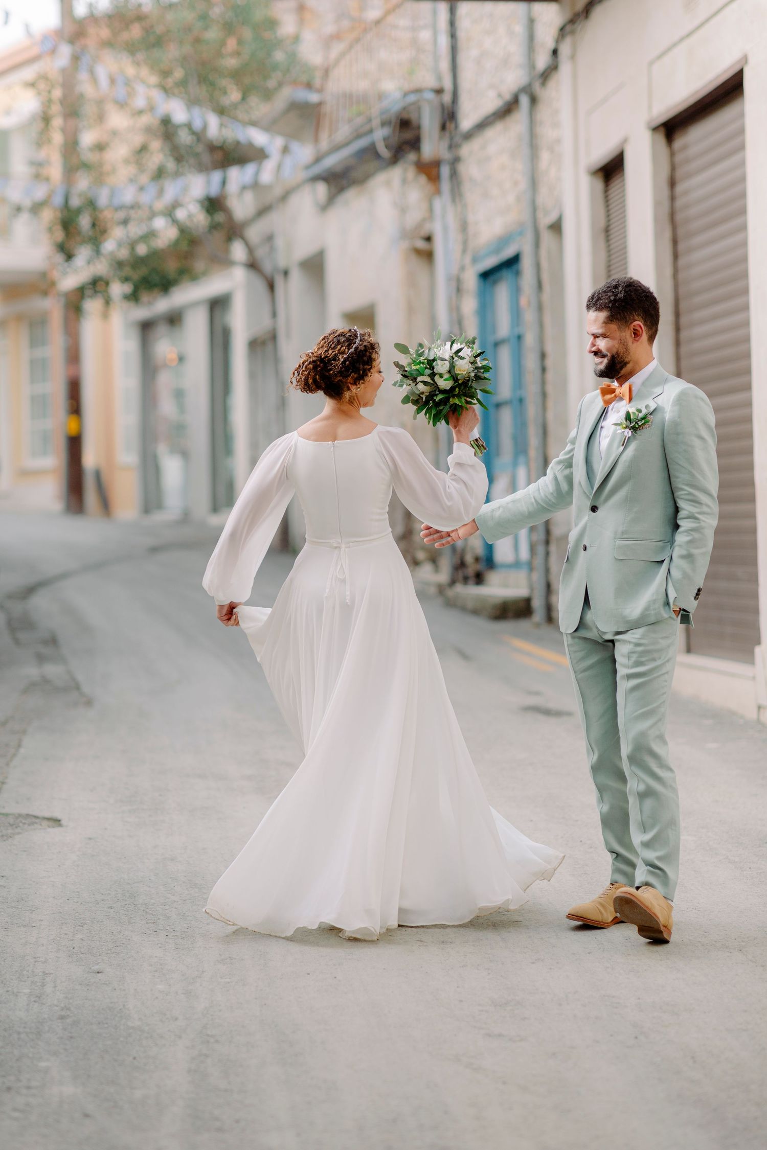 Bride in white gown and groom in mint suit hold hands walking down a charming European street on their wedding day.