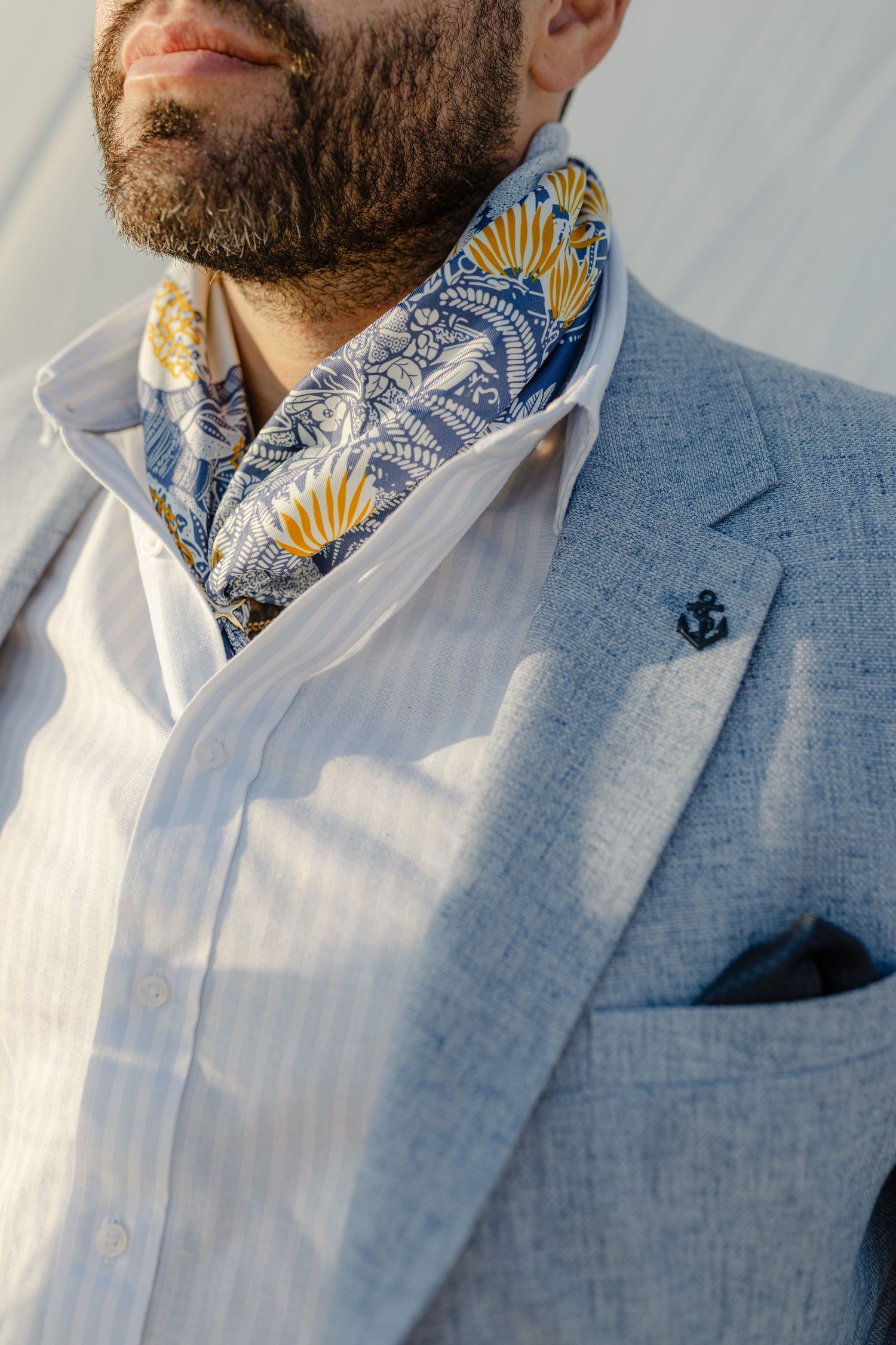 Man in light blue blazer wearing a floral blue and yellow scarf with an anchor lapel pin.