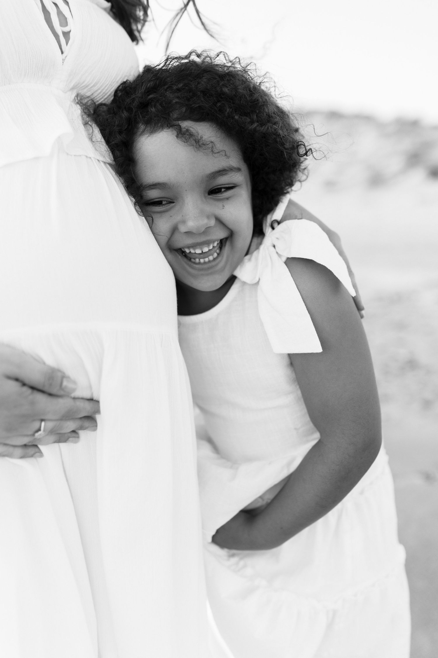 Black and white photo of a joyful young girl in white dress hugging pregnant mothers belly on a beach.