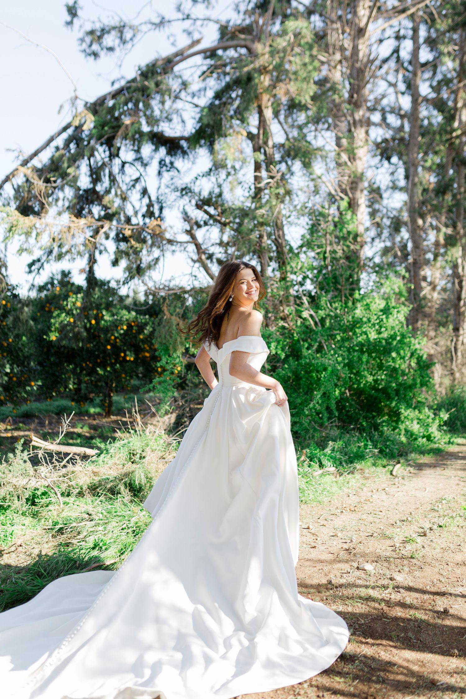 Bride in elegant off-shoulder white gown with long train poses gracefully on a sunlit forest path.