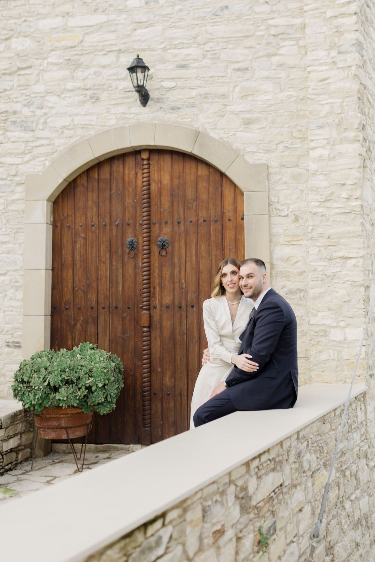 Couple in elegant attire sitting together in front of a rustic wooden arched door on a stone building.