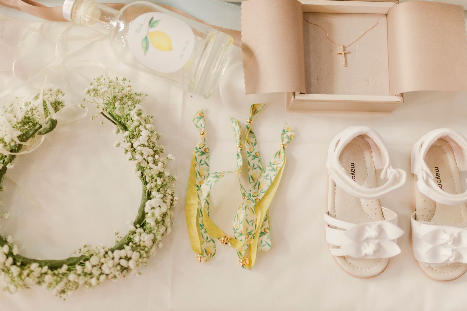 Flat lay of flower girl accessories including a baby's breath crown, white sandals, gold hair clips, and a cross necklace.