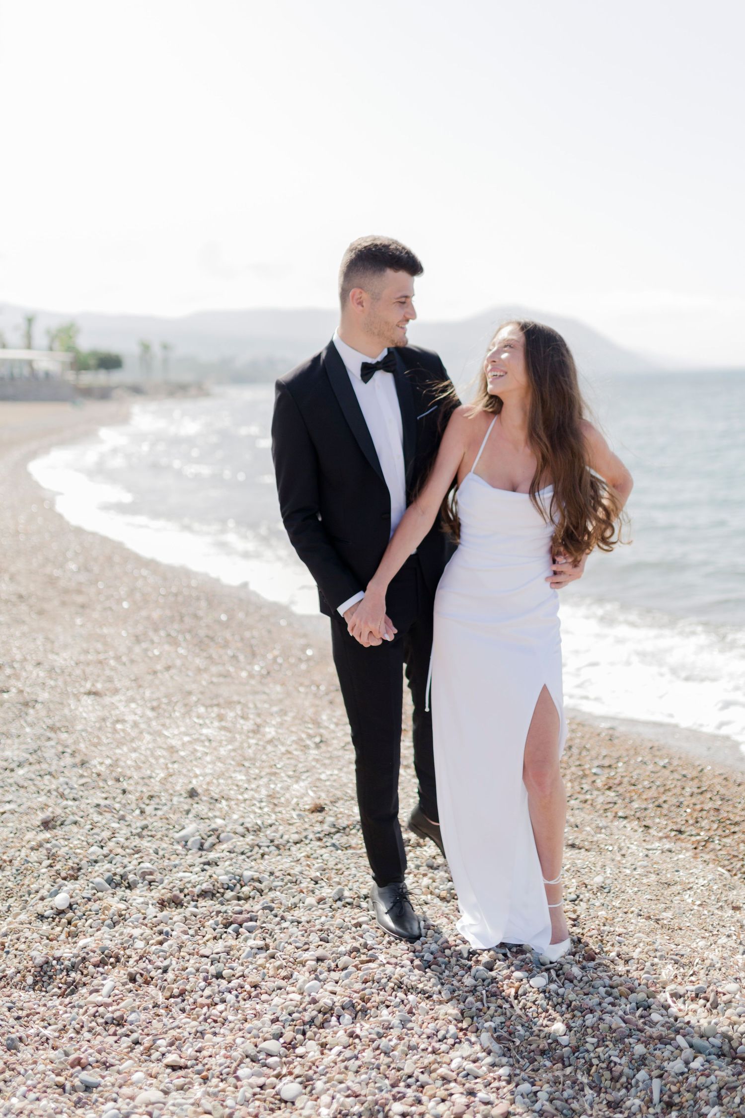 Bride in white slit gown and groom in black tuxedo walking hand in hand on a sunny pebble beach.