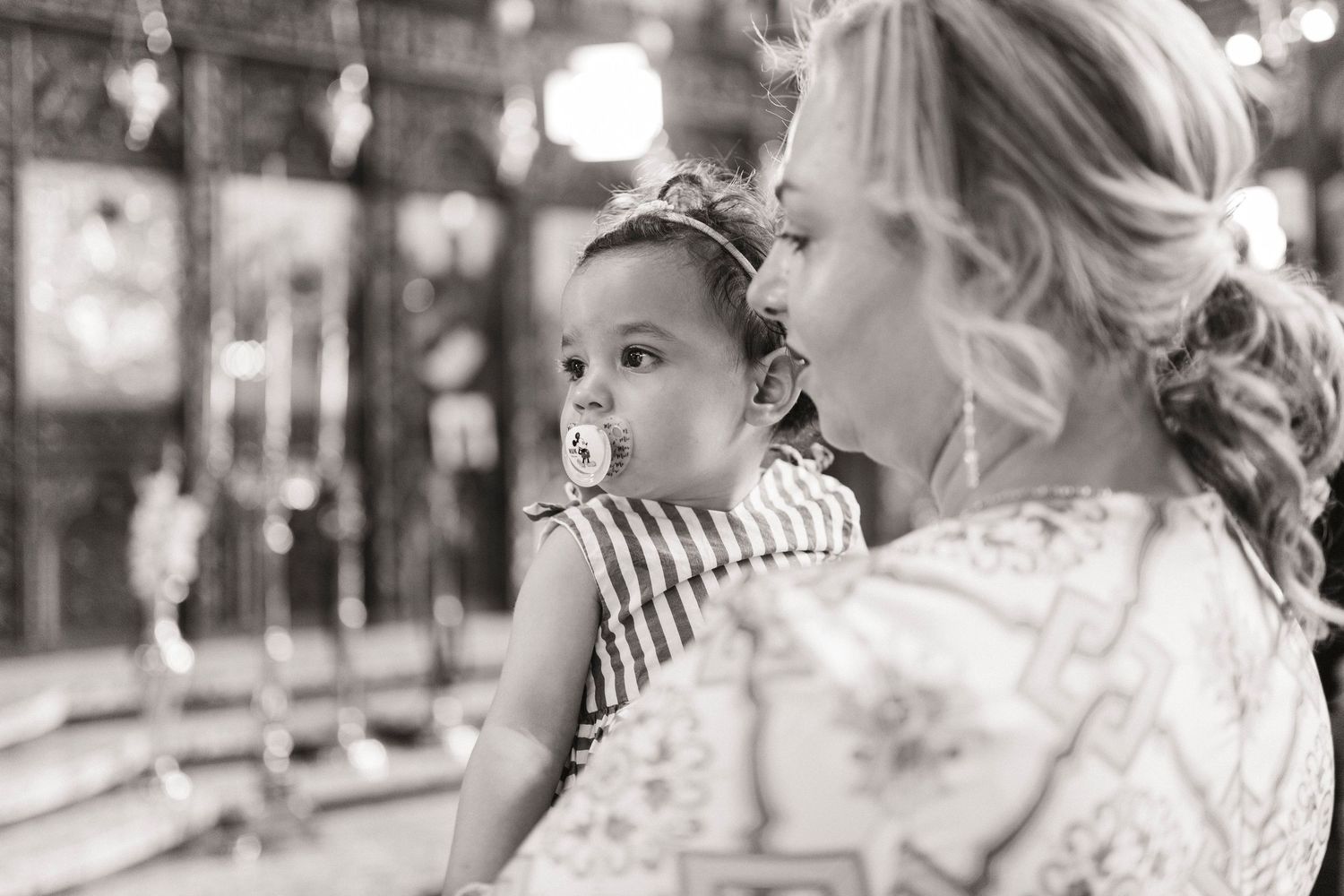 Black and white photo of a woman holding a baby with a pacifier inside a church during a baptism ceremony.