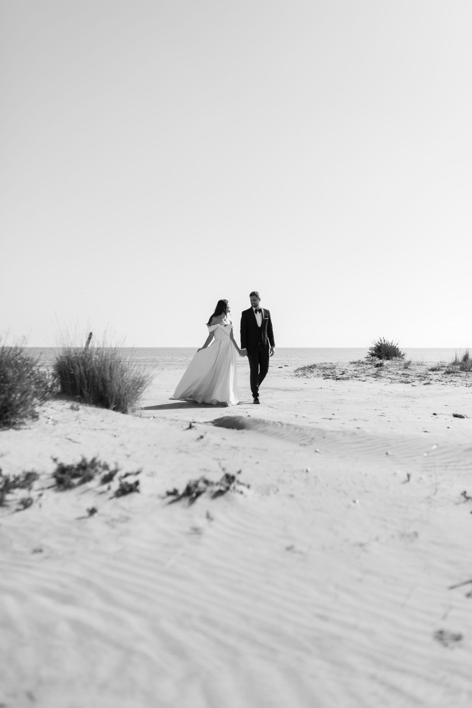 Black and white photo of bride and groom holding hands walking on sandy beach dunes toward the ocean.