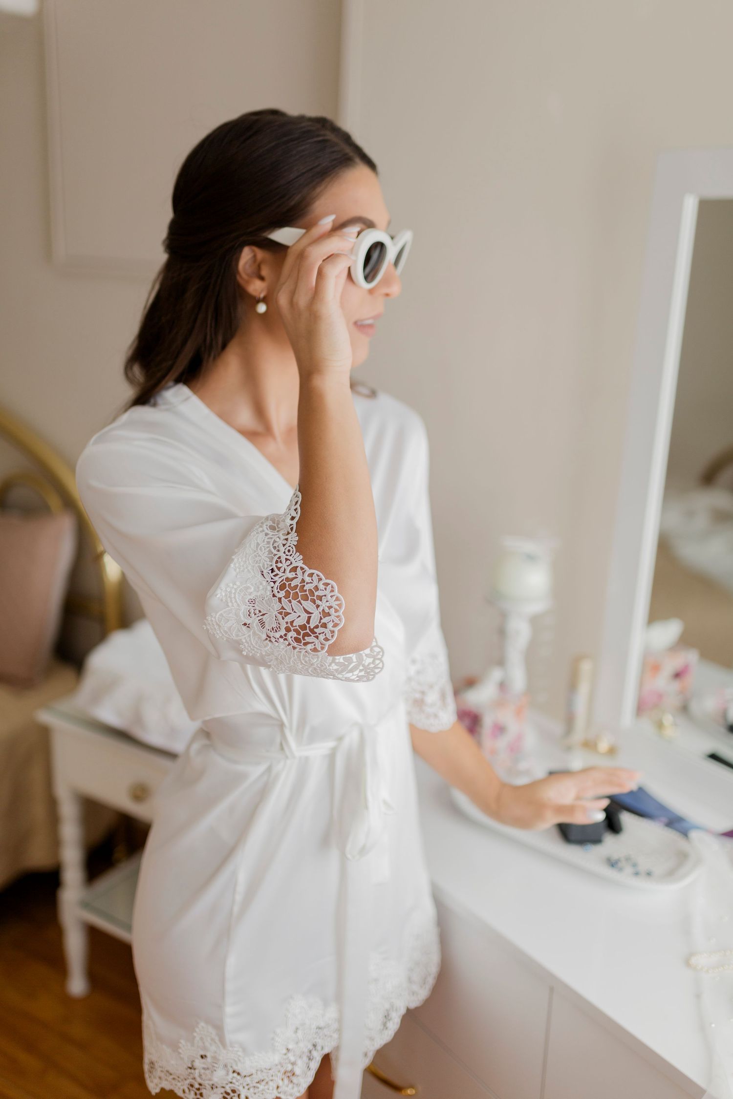 Bride in white lace robe trying on white cat-eye sunglasses at a vanity mirror while getting ready.