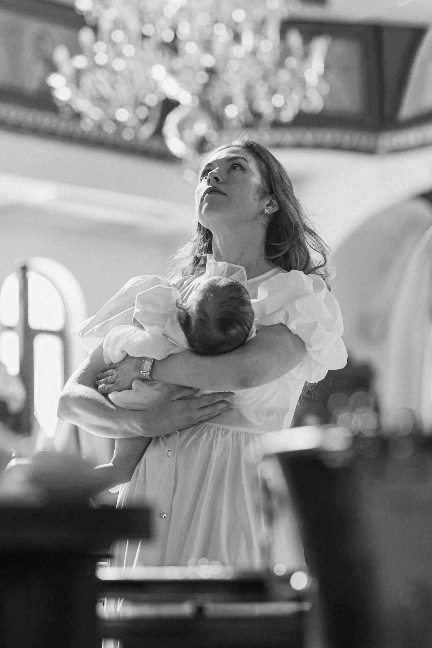 Black and white photo of a woman holding a baby in a church, looking up at a chandelier.