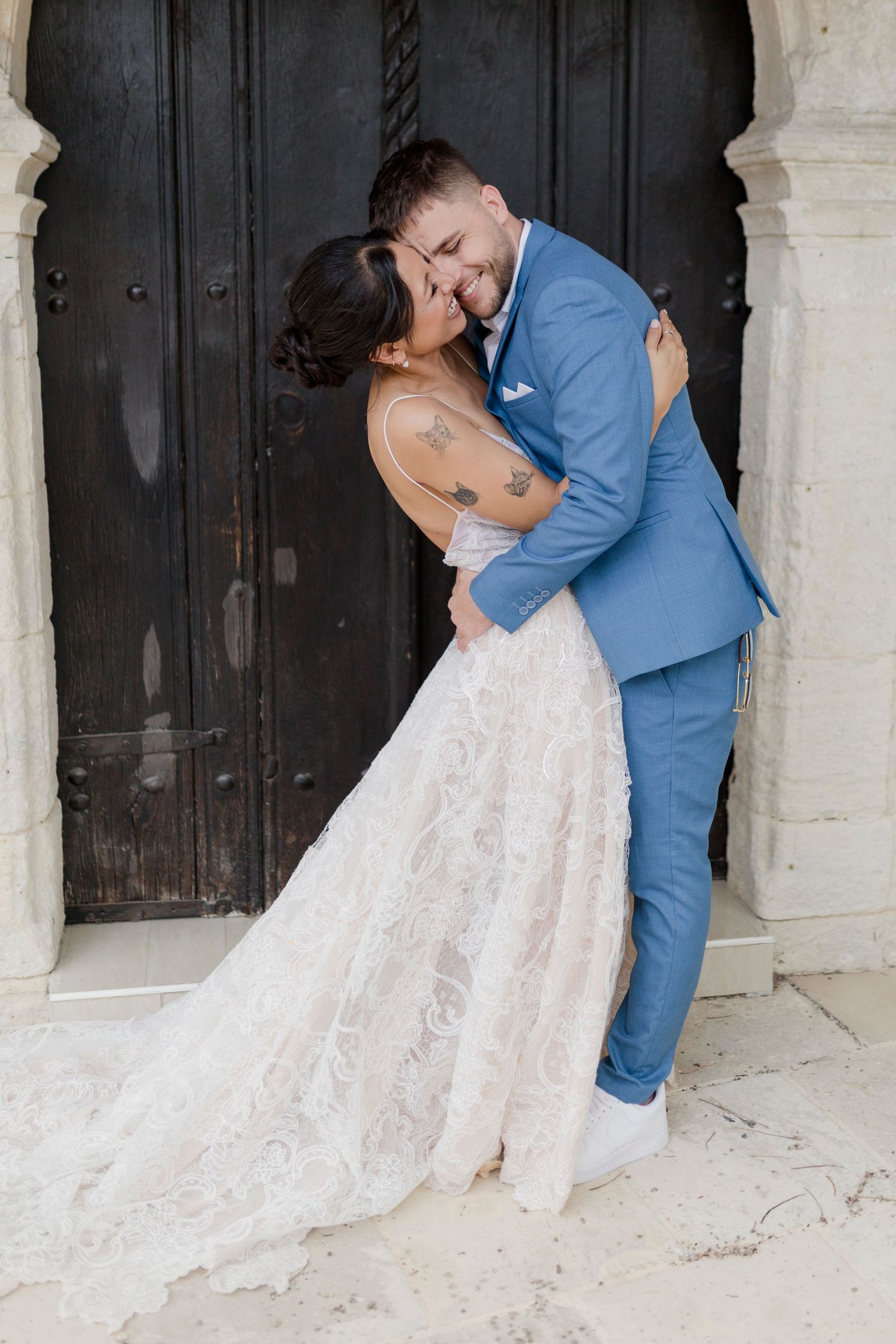 Bride and groom share romantic embrace in front of ornate dark wooden door, bride in lace gown.