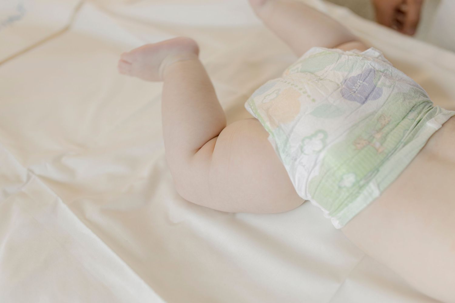 Baby lying on white bed wearing a colorful disposable diaper, showing chubby legs and bottom.