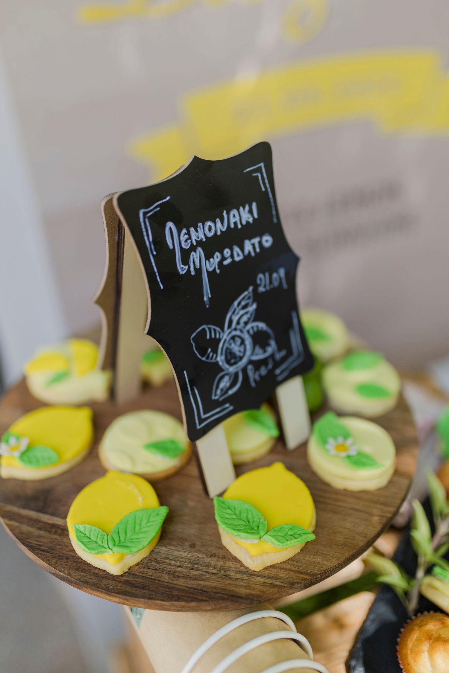 Lemon-decorated sugar cookies on wooden stand with chalkboard sign reading Lemonaki Mpompato at party.