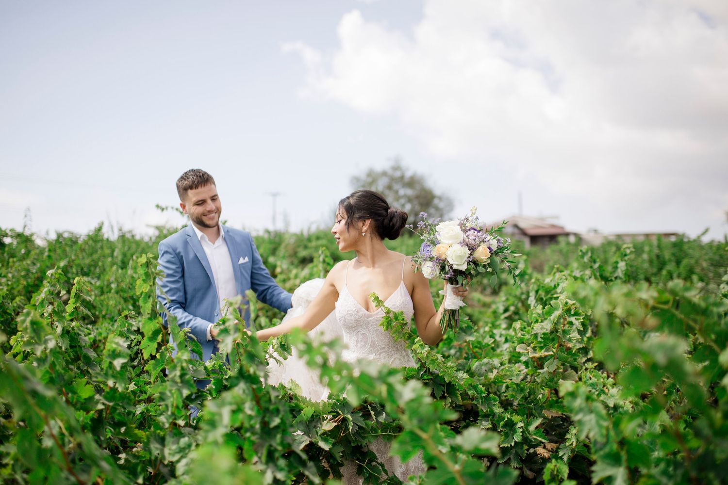 Bride and groom walking through lush vineyard, bride holding bouquet, groom in blue suit, on their wedding day.