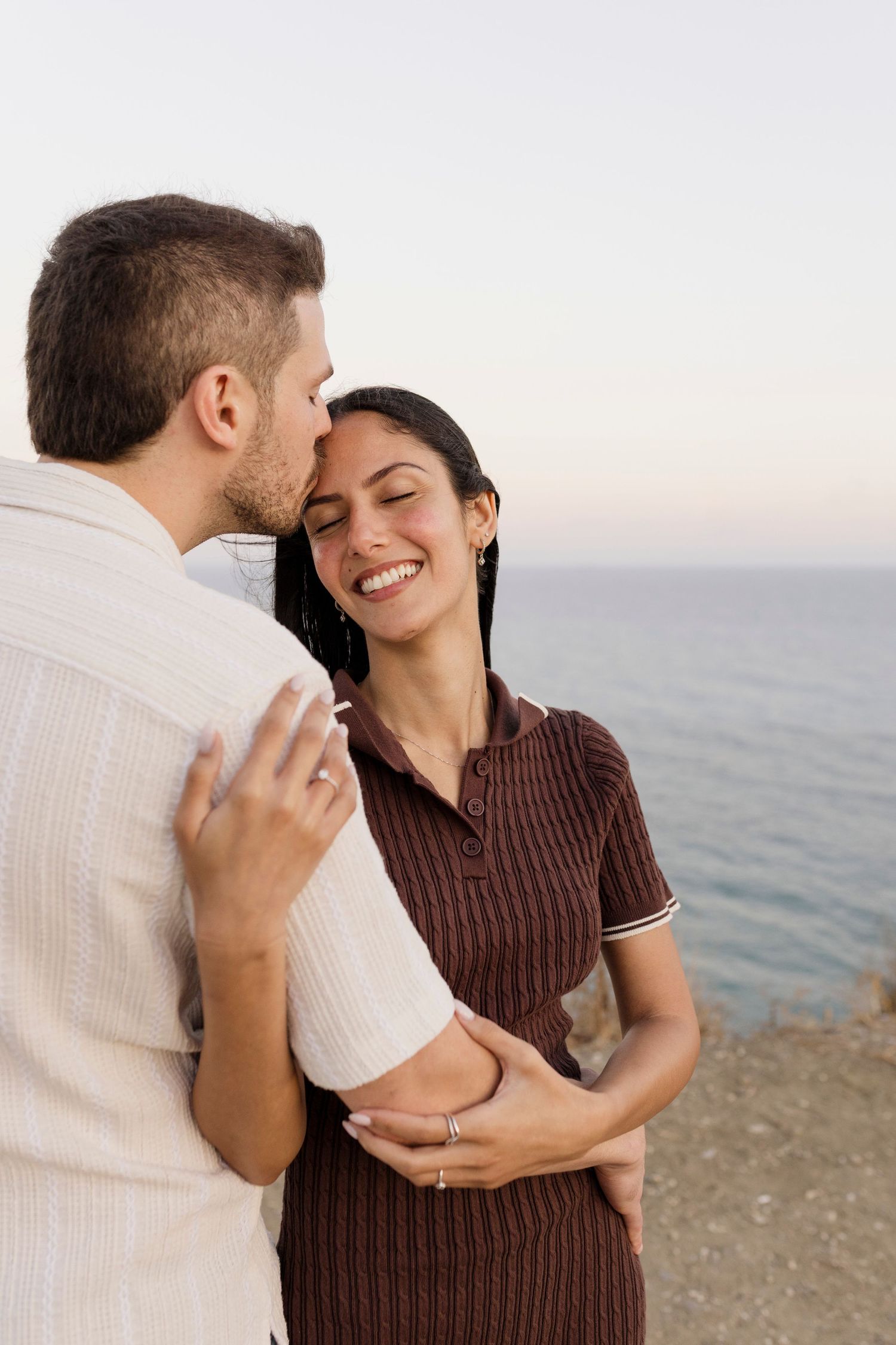 Couple embracing and sharing a tender moment by the ocean at dusk, woman smiling joyfully in brown dress.