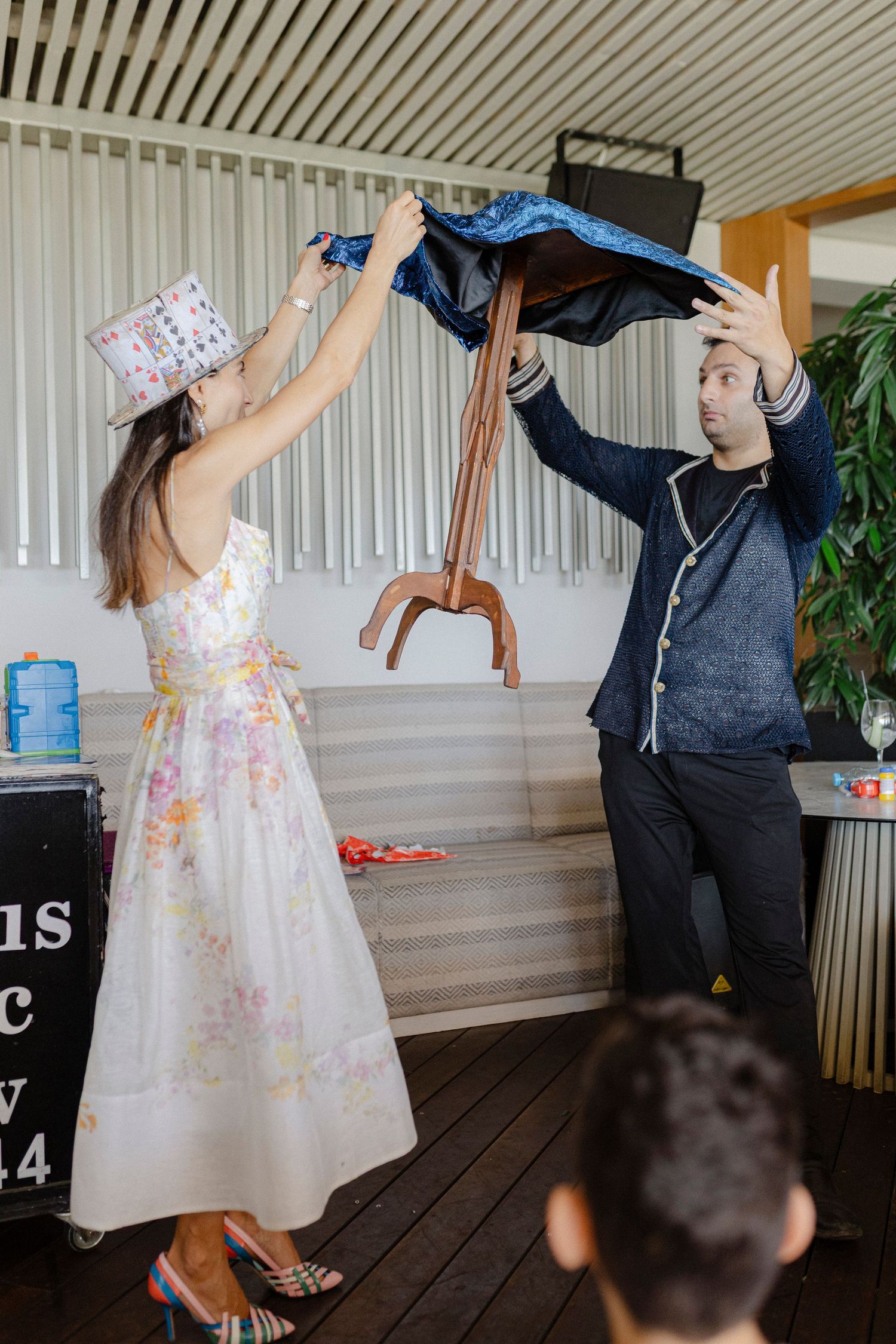 A woman in a floral dress and a man in a denim jacket lift a chair together at an indoor event.