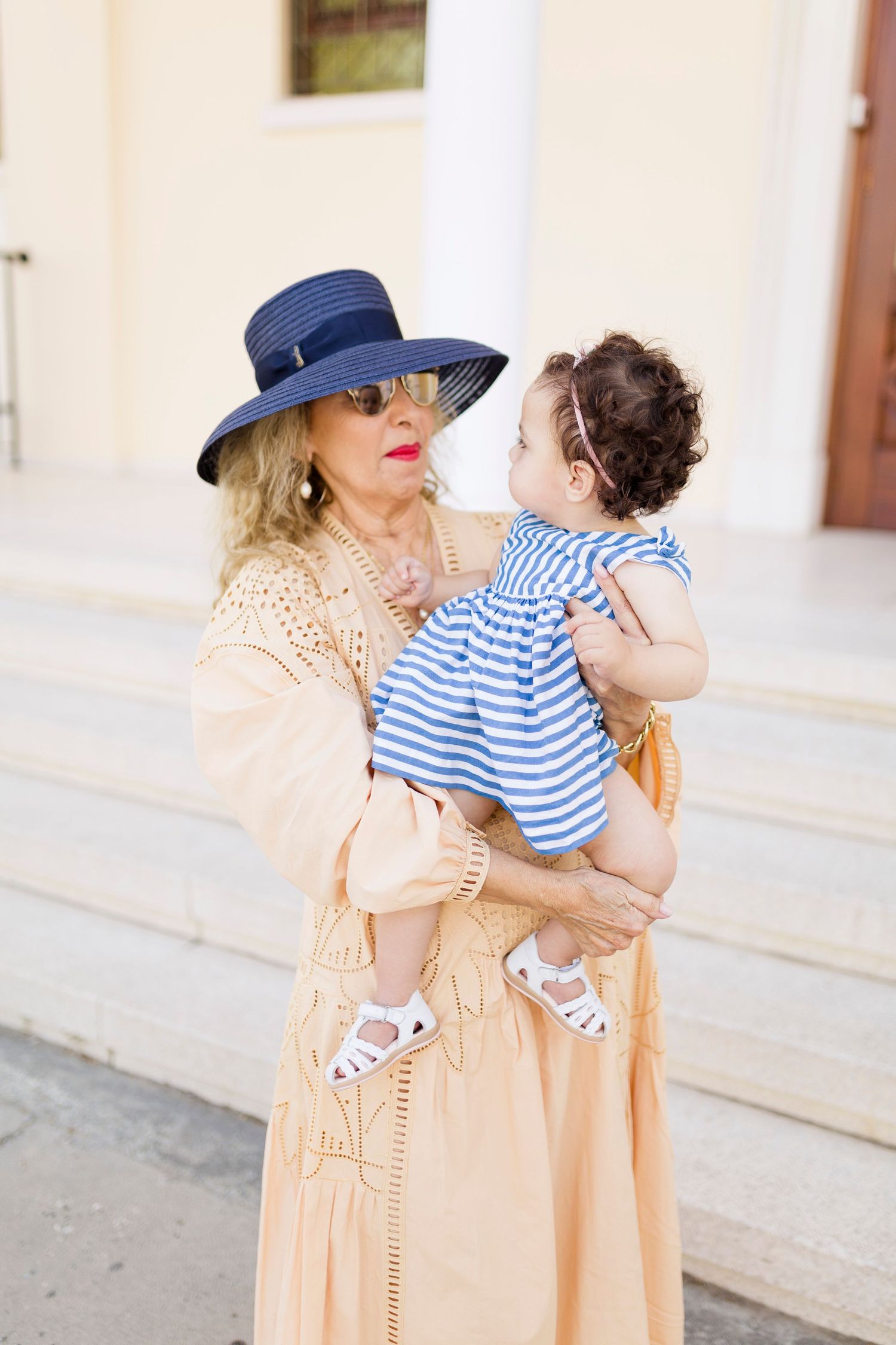 Elegant grandmother in blue sun hat and peach dress holding a baby in blue striped outfit on sunny steps.
