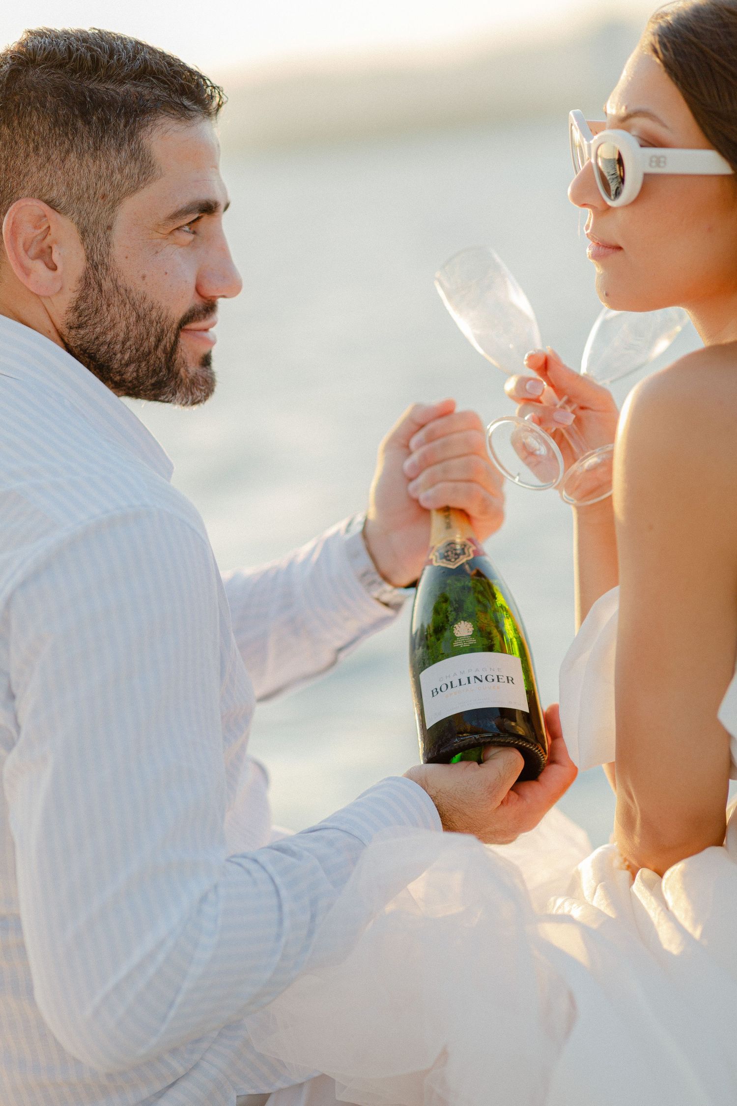 Couple sharing Bollinger champagne on a beach, dressed in white outfits, celebrating a romantic occasion.