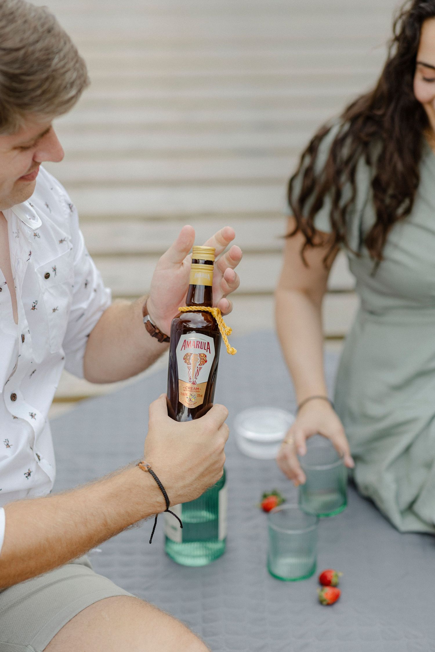 Man opening Amarula bottle while woman prepares green glasses with strawberries at an outdoor gathering.