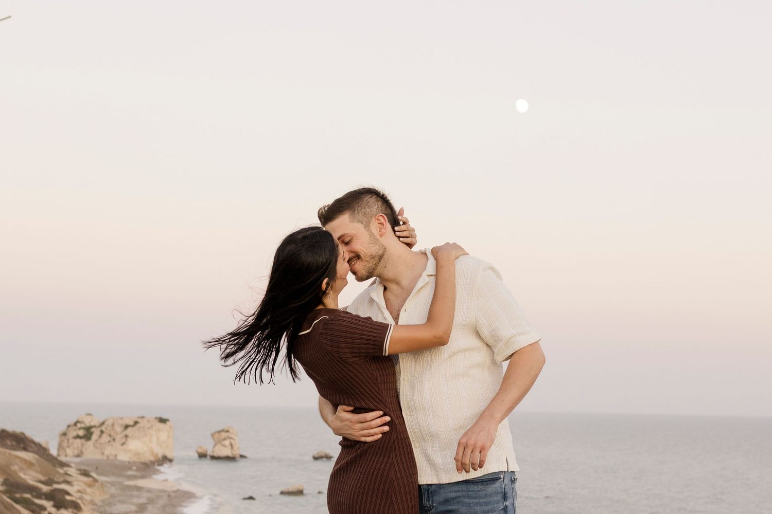 Romantic couple embracing and kissing near ocean rocks at sunset with moon visible in pastel sky.