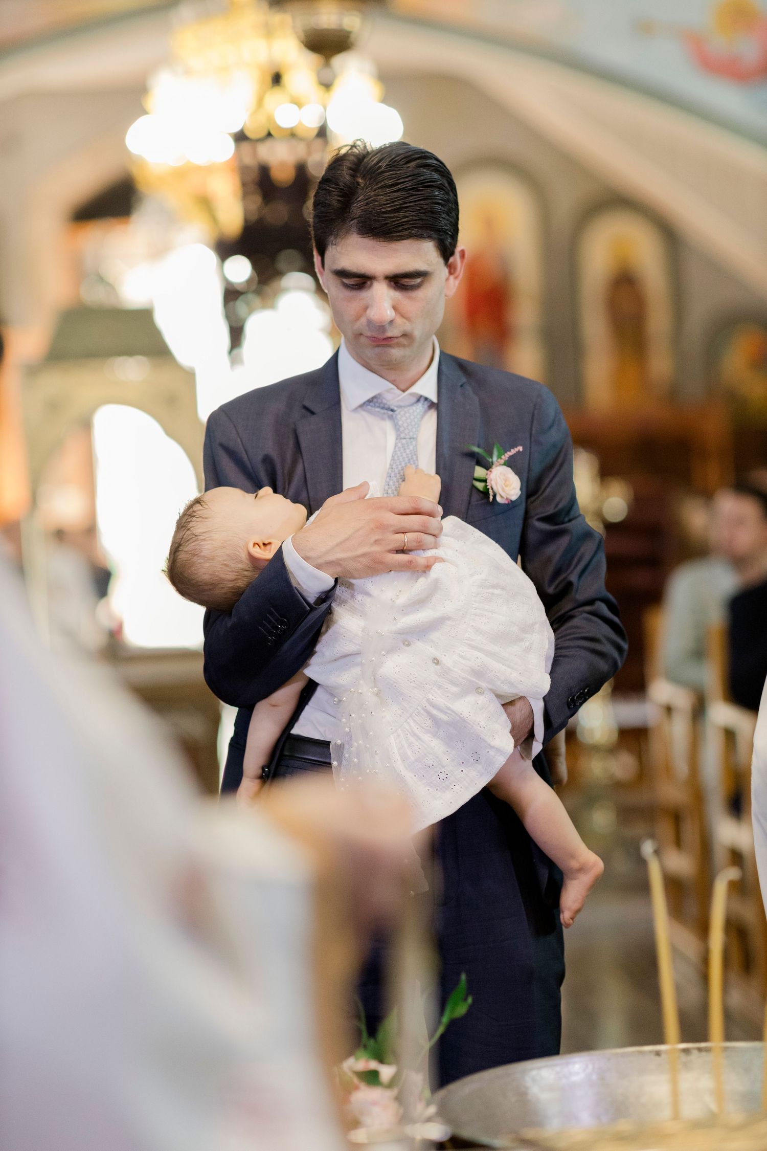Man in suit holding infant during Orthodox church baptism ceremony with chandelier and icons in background.