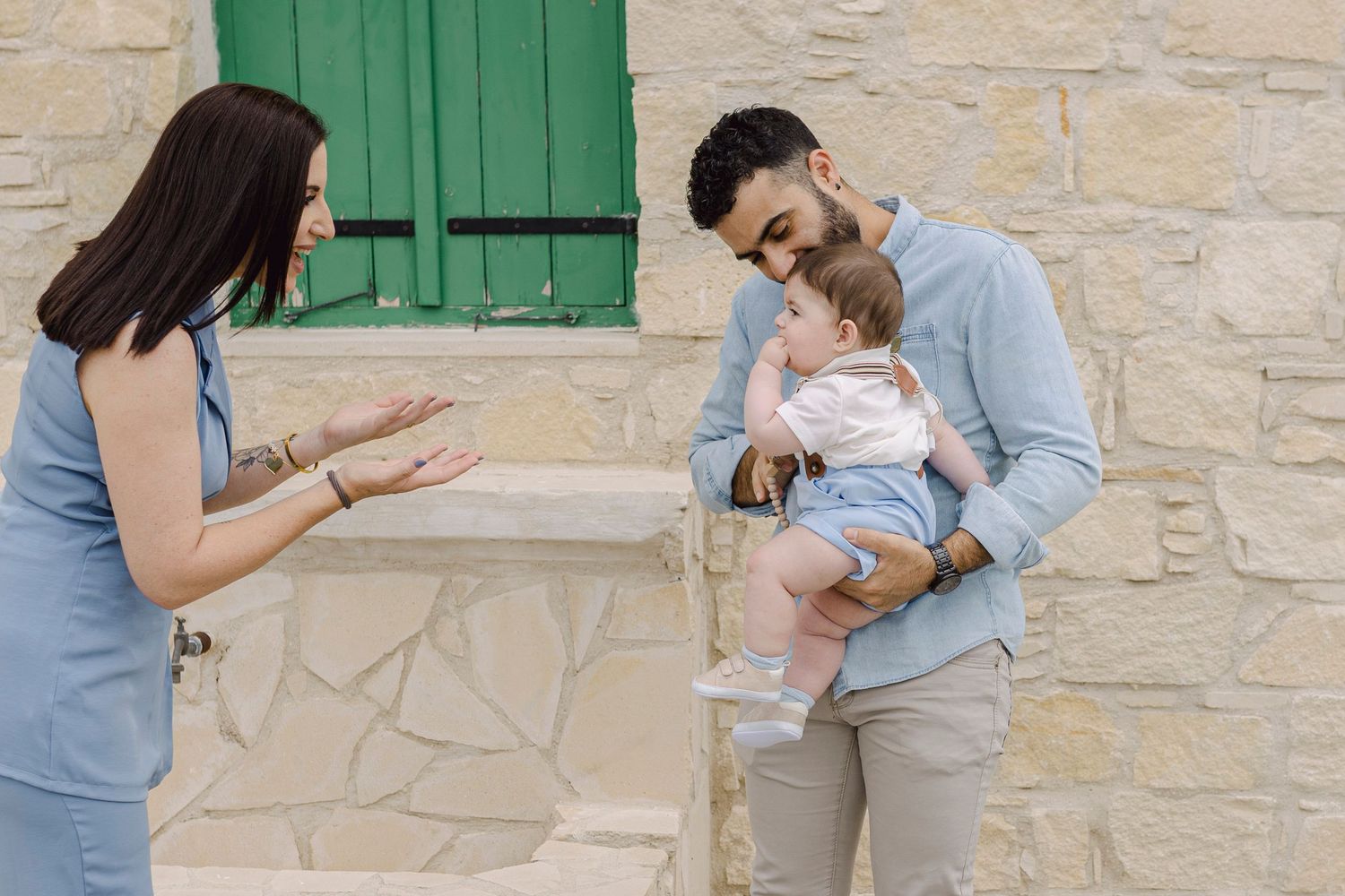 Young family with mother, father holding baby outdoors near stone wall with green window shutters.