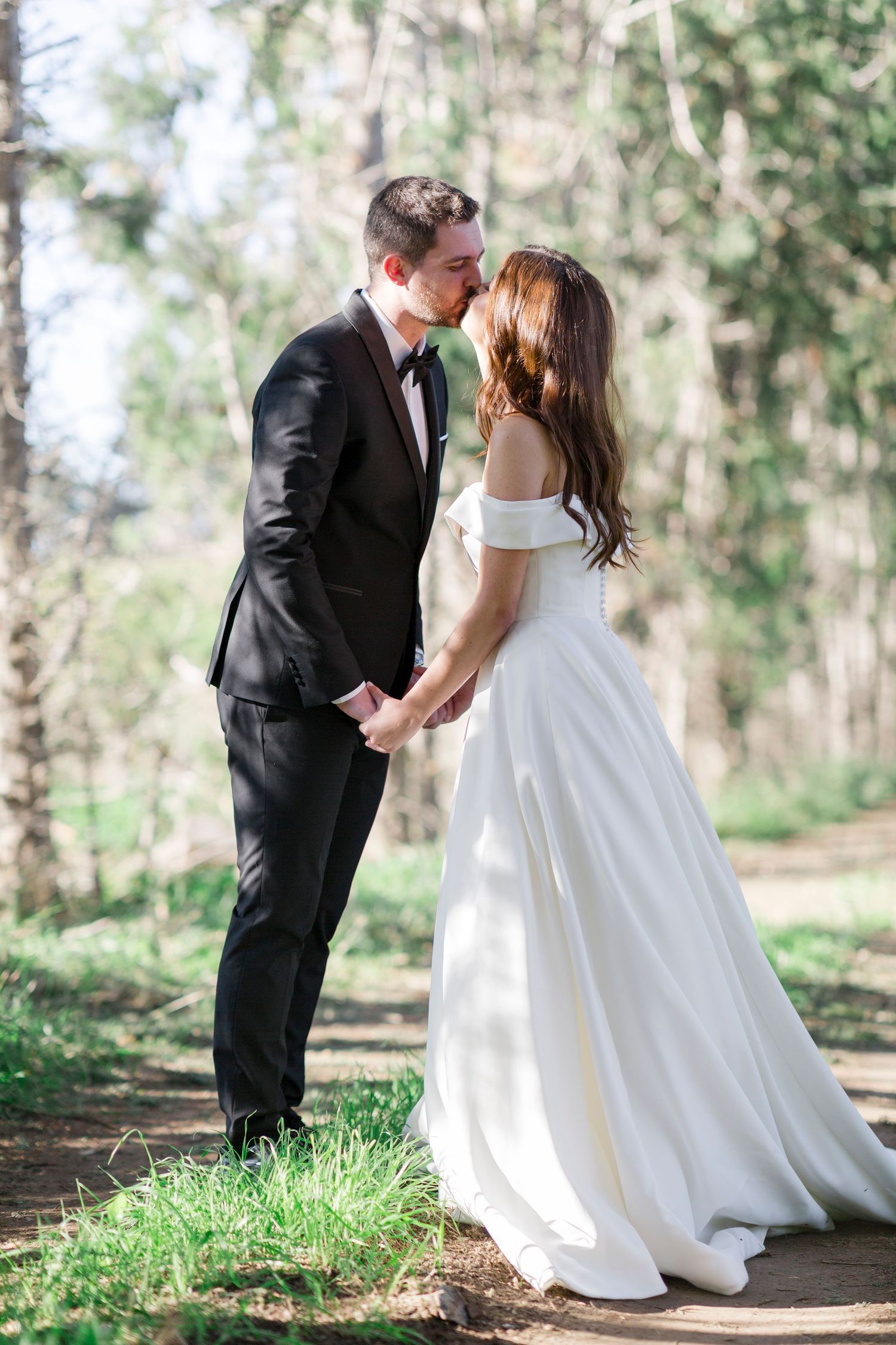 A bride and groom share a kiss while holding hands in a sunlit forest, she in a white off-shoulder gown, he in a black tuxedo.