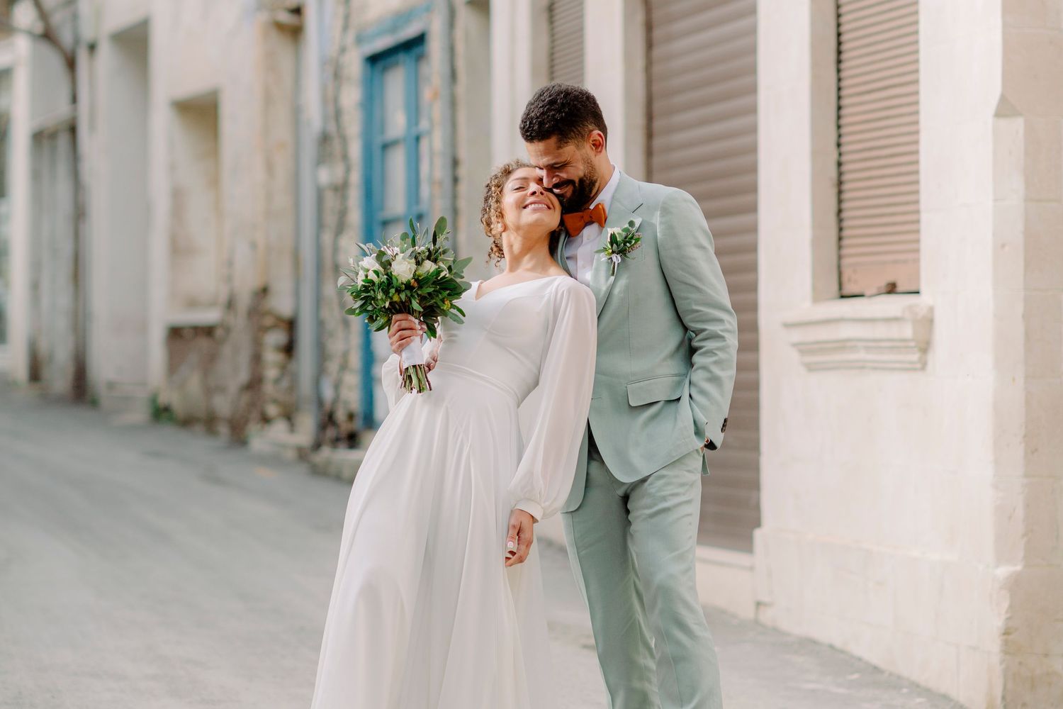 A smiling bride in a white gown holding a bouquet walks with a groom in a light blue suit on a charming European street.