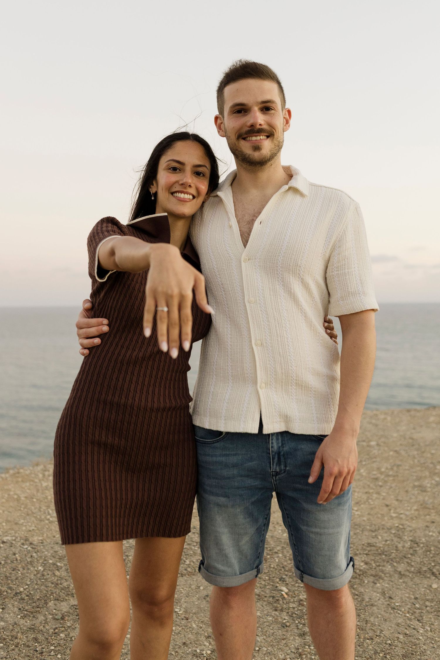 A smiling couple embraces by the sea at sunset, the woman showing off her engagement ring.